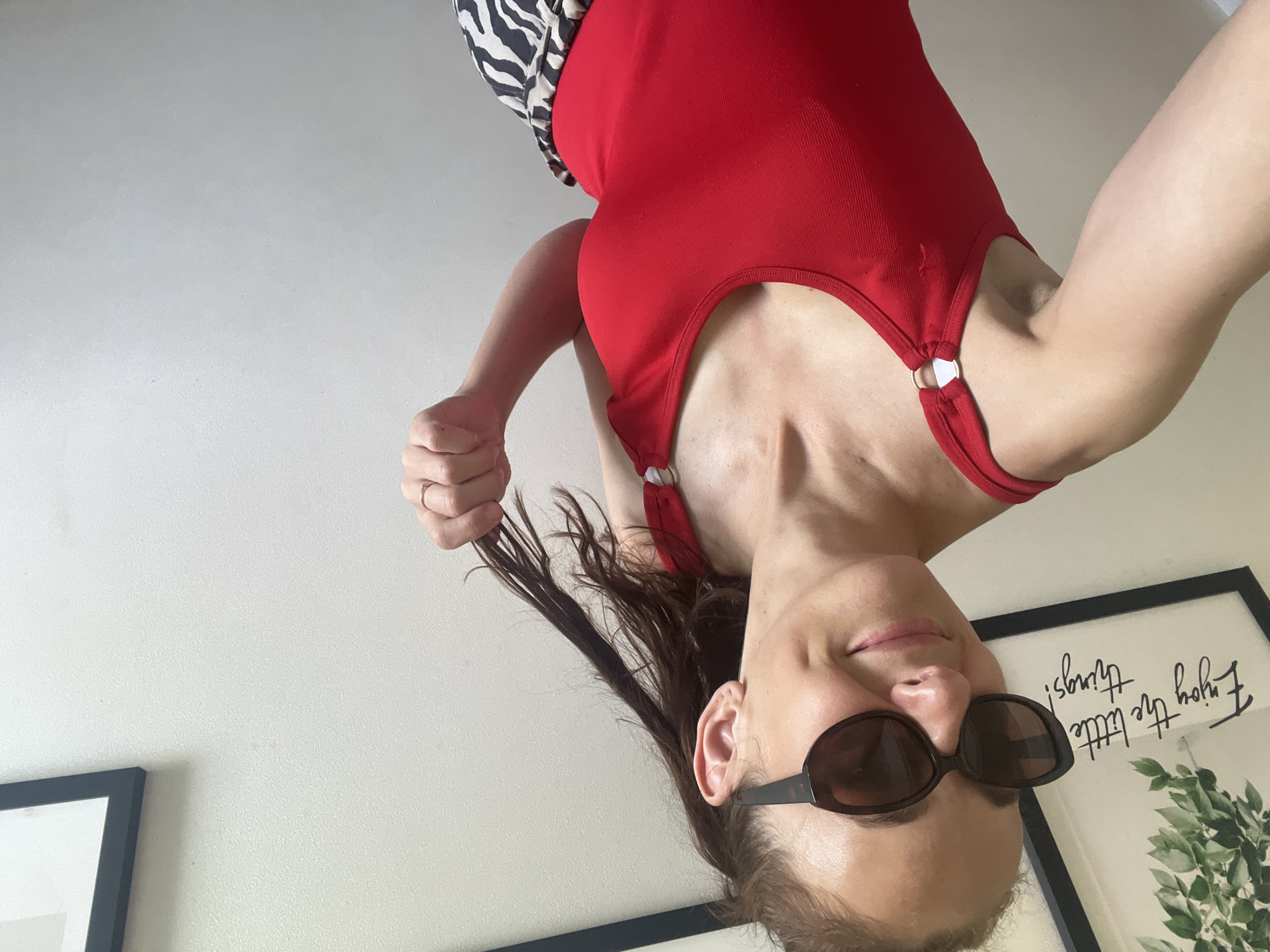 The image shows a woman indoors, with a white wall as the background. She is wearing a red tank top and what appears to be a black and white patterned garment on her left shoulder. She has sunglasses on and is holding a strand of her dark hair. Her face is tilted downwards, and she is smiling slightly. Two framed pictures are visible on the wall behind her. One has a black frame and appears to contain text and an image of greenery. The visible text reads "Enjoy the little things!". The other frame is also black. The lighting suggests it is daytime.