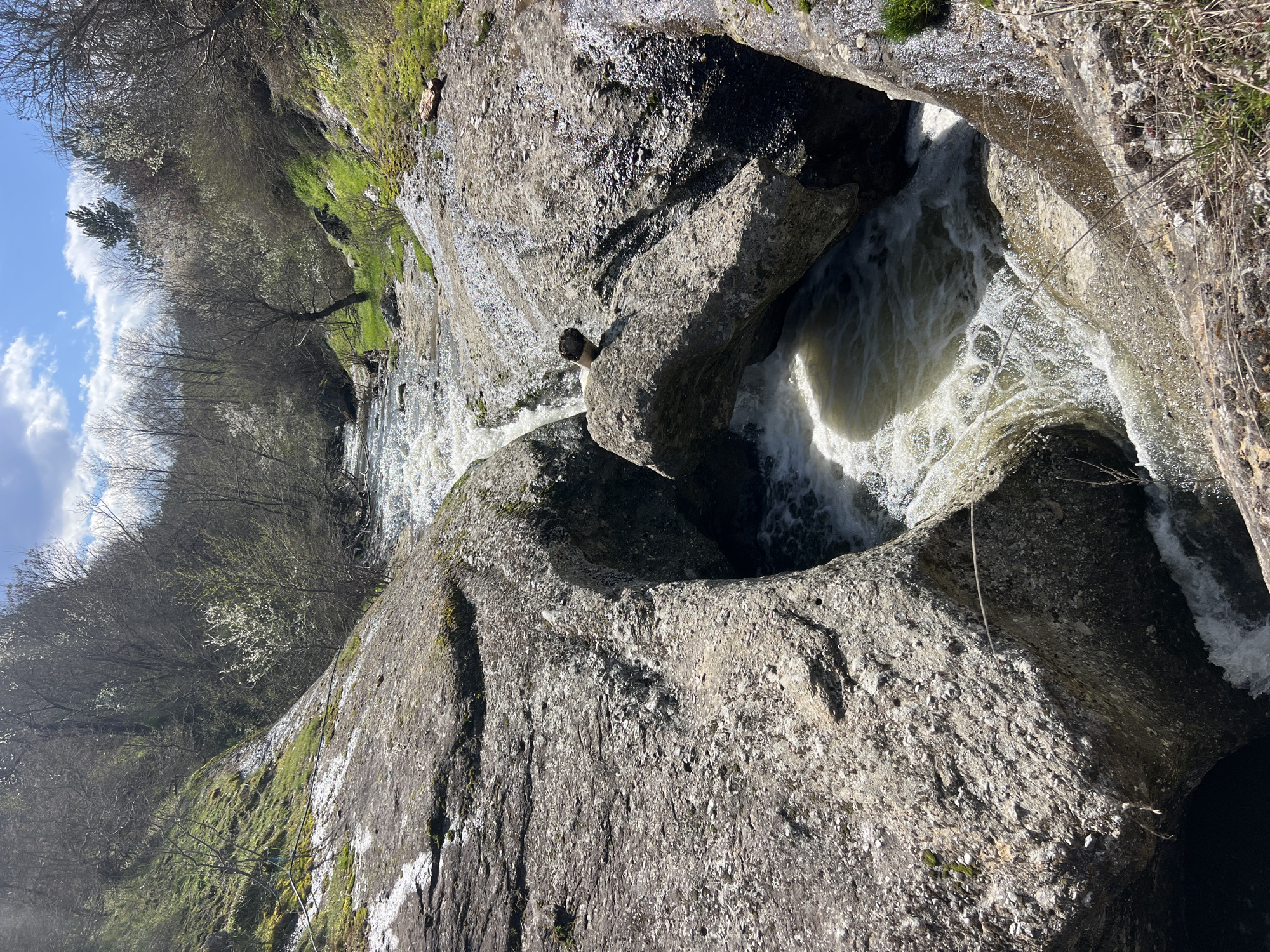 The image shows a cascade of water flowing through a rocky landscape. The location is identified as the city of Suhovo, Bulgaria. The water appears turbulent and foamy as it rushes between and over the gray, moss-covered rocks. The rocks exhibit varied textures and shapes. In the background, trees and grass are visible on the banks of the watercourse. The sky is blue with some cloud cover. The perspective is from above, looking down at the cascade.