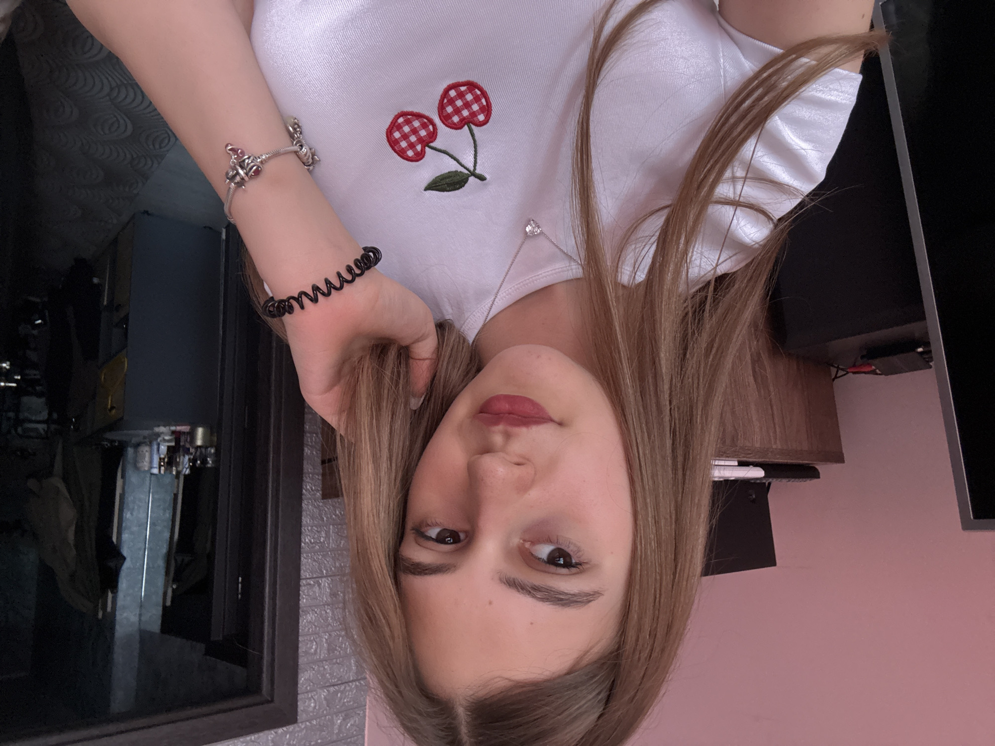 A young woman is taking a selfie, the camera is rotated 180 degrees. She is wearing a white shirt with a cherry patch and a silver necklace with a stone. She has several bracelets on her left wrist, her left hand is by her face, touching her hair. The background includes a window, a TV, and a wall. The location in the city of Panagyurishte, Bulgaria cannot be determined from the image.