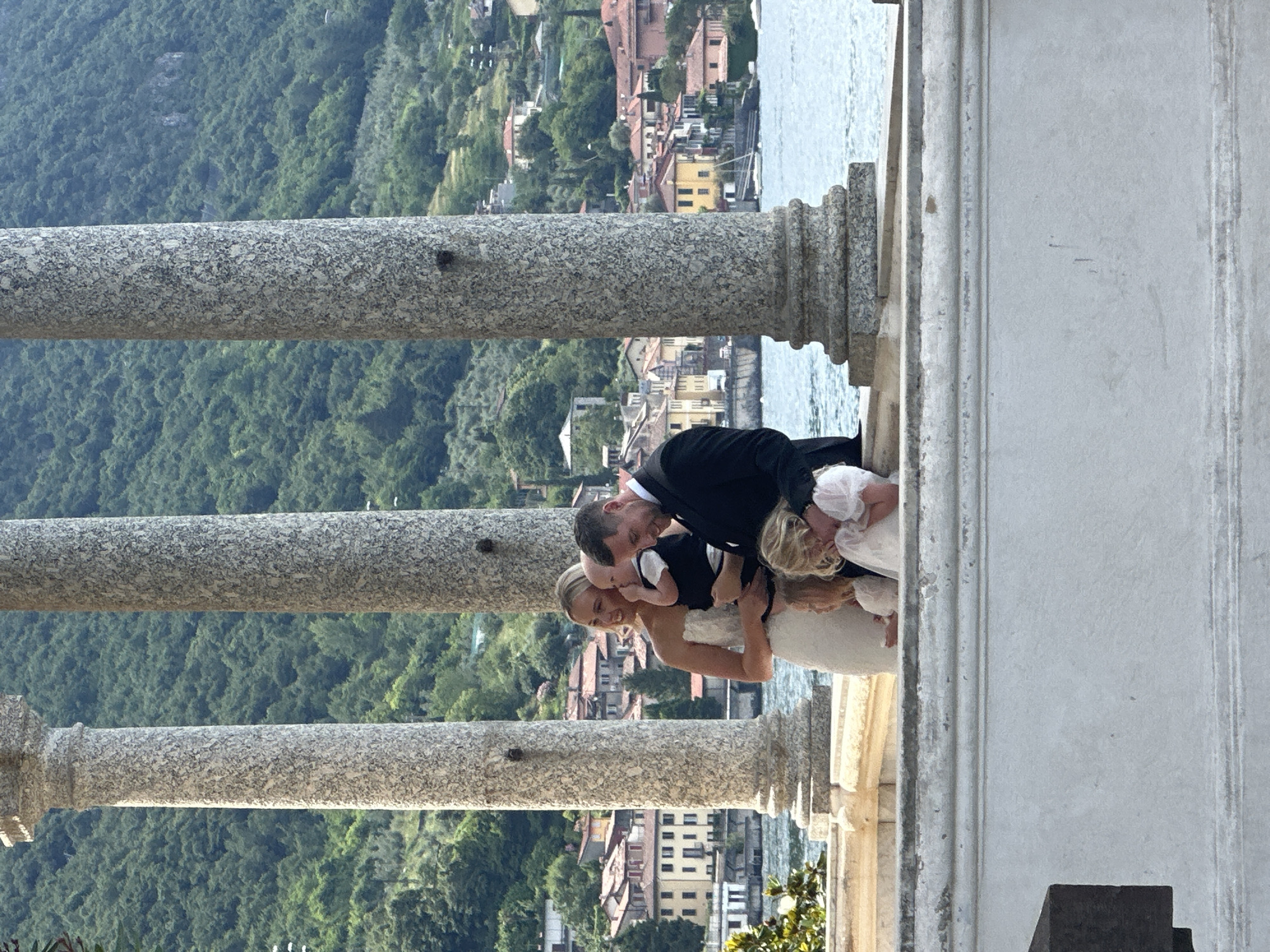 A wedding party of five is positioned on a balcony with a lake and hillside town in the background. The group consists of a bride in a white strapless dress, a groom in a black suit, a baby being held by the groom, and two young girls. The baby is wearing a black vest and white shirt. Both girls are wearing white dresses. The bride and groom are smiling. The two girls are looking down, with one of them hugging the groom's leg. In the background, there is a body of water, a town with red-tiled roofs and buildings on a green, tree-covered hillside. The scene is framed by four large, fluted stone columns. The overall impression is of a happy family occasion in a picturesque Italian setting.