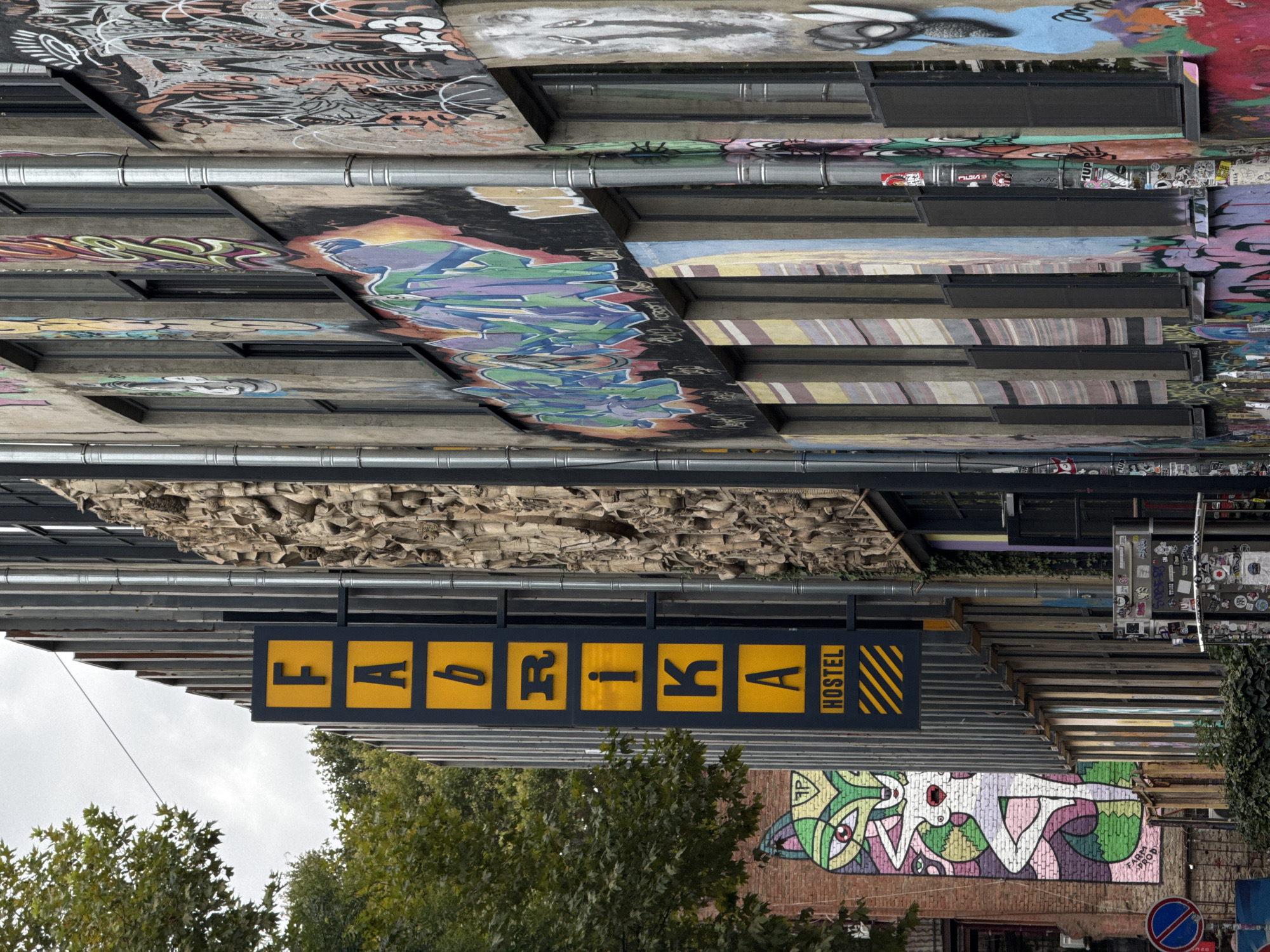 This is a street-level, eye-level, medium shot of the exterior of a building covered in graffiti in Tbilisi, Georgia. The building appears to be a hostel, as indicated by a vertical yellow sign with black lettering spelling out "FABRIKA HOSTEL". The building's facade is a mixture of concrete, metal, and exposed brick, adorned with various graffiti art pieces. On the left side of the image, a tall, colorful mural of a stylized fox-like creature is visible on a brick wall. The main building features a textured, relief sculpture on its upper section, above rows of windows. Various pipes and conduits run vertically along the building's exterior. In the foreground, there is a low-lying area with dense green foliage, and to the right, a metal structure covered in stickers. The sky is overcast, suggesting a cloudy day.
