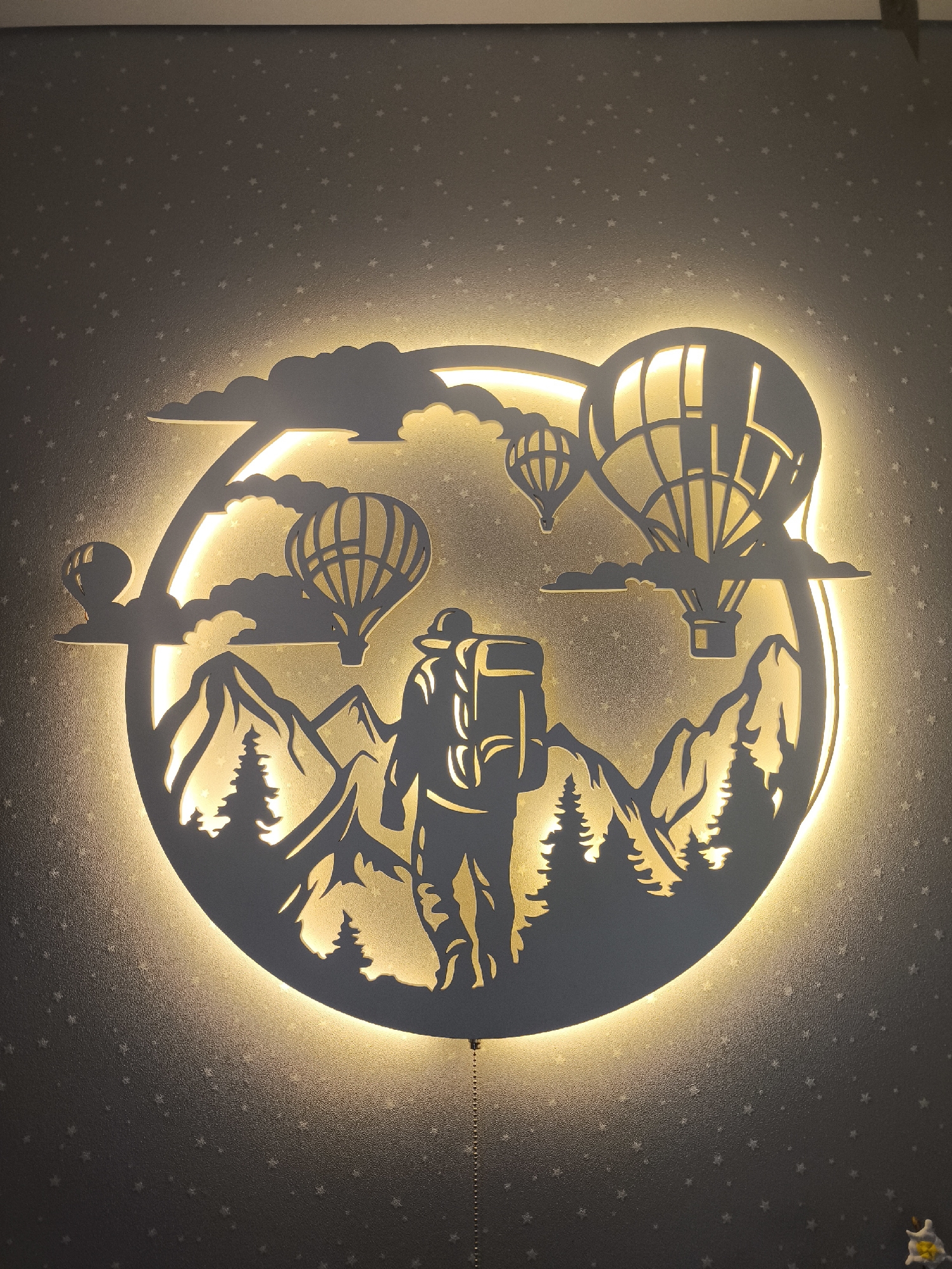 An illuminated, circular, decorative wall fixture is centered in the image against a speckled gray wall. The fixture is a silhouette cut-out depicting a hiker with a backpack in a mountain landscape, with hot air balloons floating above. Clouds are also silhouetted in the design. The fixture emits a warm, yellow light from behind, highlighting the cut-out shapes. A pull chain hangs from the bottom of the fixture. The location is not apparent in the image.