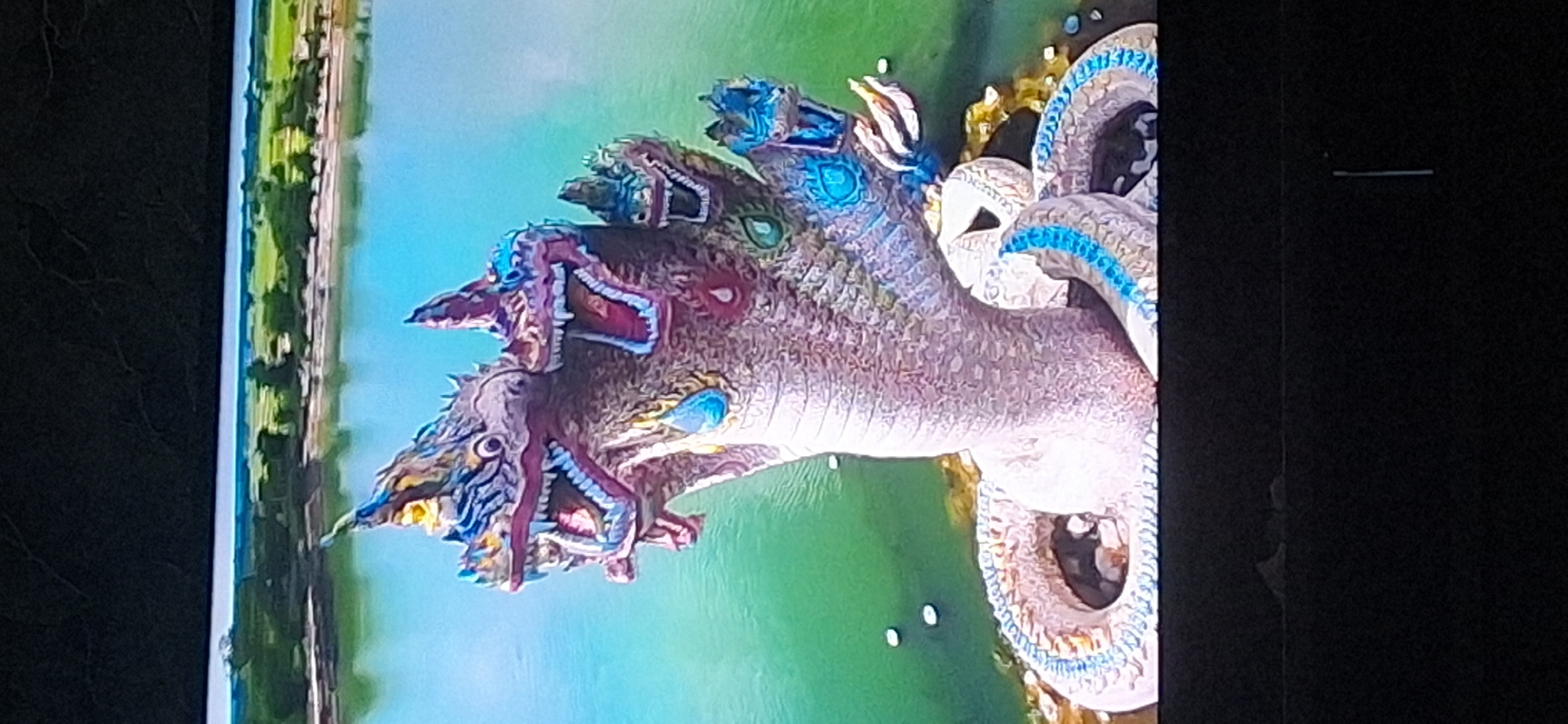 This image shows a statue of a multi-headed serpent, possibly a Naga, in a body of water. The serpent has a pinkish-grey body with intricate detailing and multiple heads, each with an open mouth and sharp teeth. The heads are adorned with colorful headdresses and have jewel-like eyes. The serpent's body is coiled and partially submerged in green water. In the background, a lush green landscape with trees and a body of water is visible under a bright sky. The bottom of the image is dark, suggesting it might be a screen or a frame. There are no people visible in the image.
This statue is located in Bila Tserkva, Ukraine.