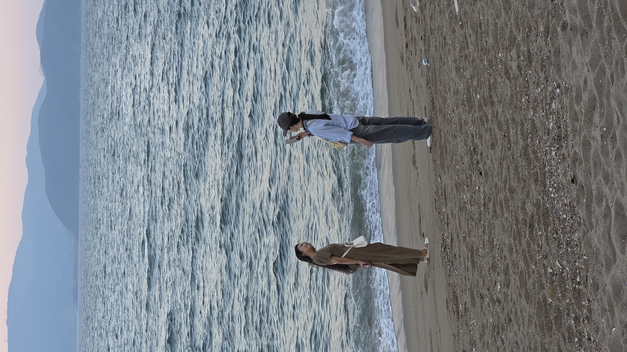 The image depicts a sandy beach scene in Nha Trang, Vietnam, during twilight. Two young adult females are present. On the left, a woman with long dark hair, wearing a long olive-green dress, white shoes, and a white crossbody bag, stands facing right. She has her eyes closed, a subtle smile, and her hands clasped behind her back, appearing to pose for a photograph. On the right, another woman with dark hair tied back, wearing a grey baseball cap, a light blue long-sleeved shirt, dark grey wide-leg pants, and white sneakers, holds a smartphone horizontally with both hands, pointing it towards the first woman, actively engaged in photography. Small waves from the dark blue-green ocean break on the shore behind them. The background features distant, layered mountains under a pale pink and blue sky. The foreground sandy beach contains footprints, scattered shells, small pebbles, and minor litter, including a white plastic cup and a blue object near the photographer.
