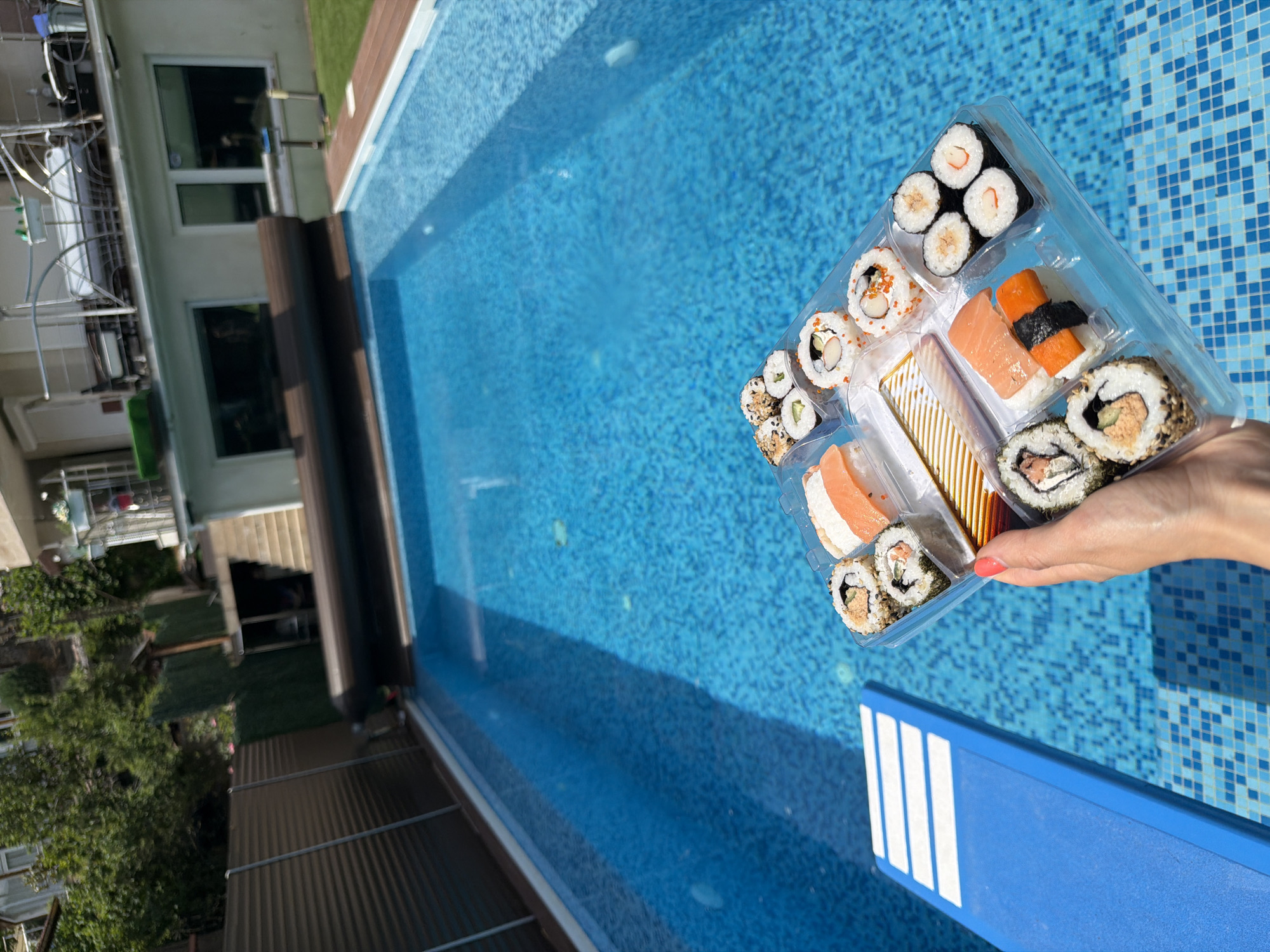 In Sofia, Bulgaria, a clear plastic container filled with a variety of sushi rolls and nigiri sits precariously close to the edge of a swimming pool. The sushi appears to be fresh, with colorful ingredients like salmon and orange-colored fillings visible. A woman's hand, with bright coral nail polish, holds the container, her fingers delicately supporting it. The swimming pool itself is a vibrant blue, its tiled bottom reflecting the bright sunlight. A blue diving board with white stripes is partially visible in the lower left corner. In the background, a multi-story building with white walls and several windows overlooks the pool area, surrounded by lush green trees and a dark corrugated fence. A dark pool cover is partially rolled up, extending across the water's surface. The overall scene captures a moment of leisure, with the inviting pool and a delectable sushi platter suggesting a relaxing day outdoors.