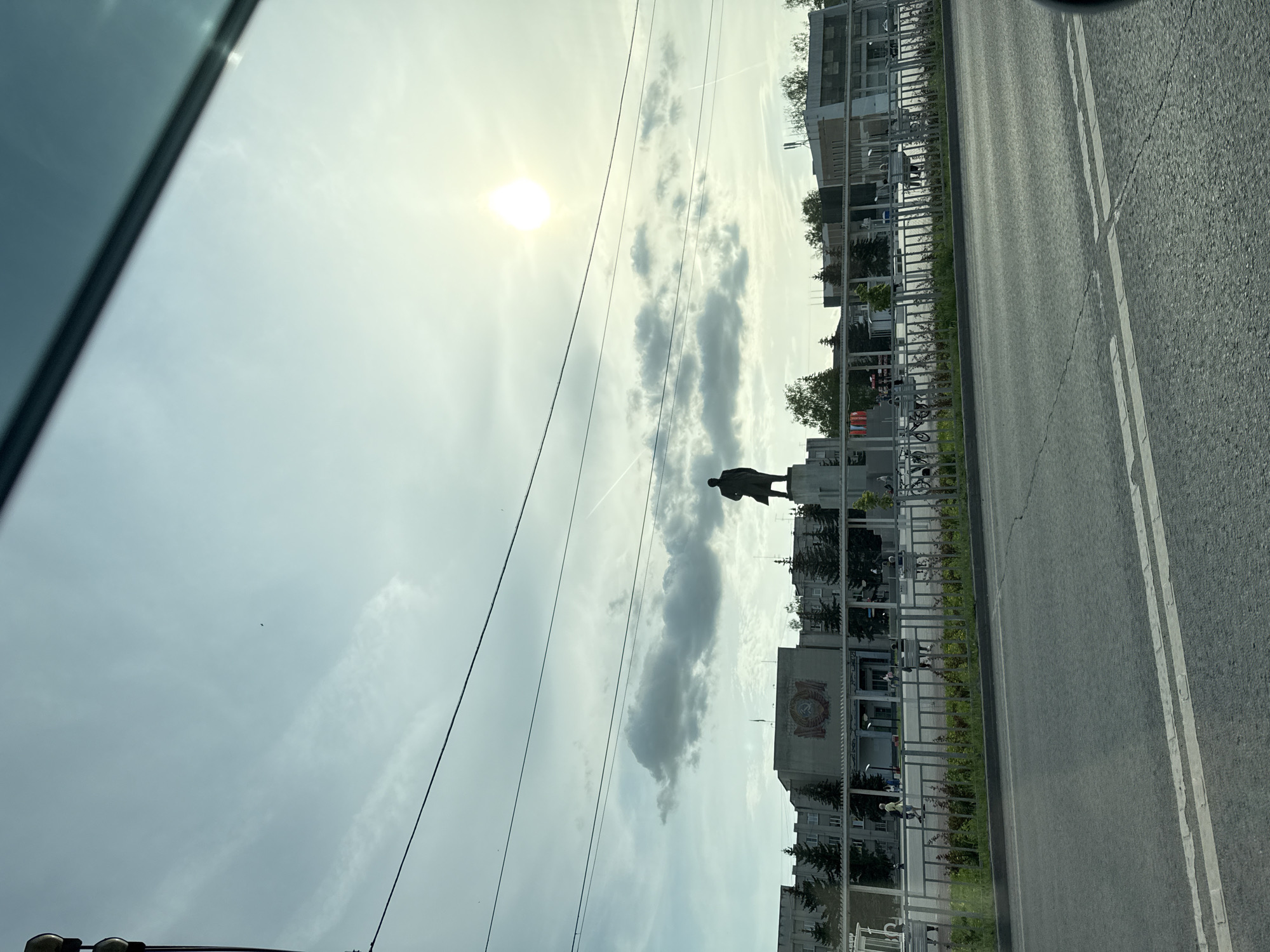 A street scene in Kstovo, Russia, includes a statue of Vladimir Lenin on a pedestal. It is partially obscured by clouds. The road is marked with white lines, and buildings with trees can be seen alongside it. The sky is bright, with the sun visible through the clouds. The photo was taken from inside a car.