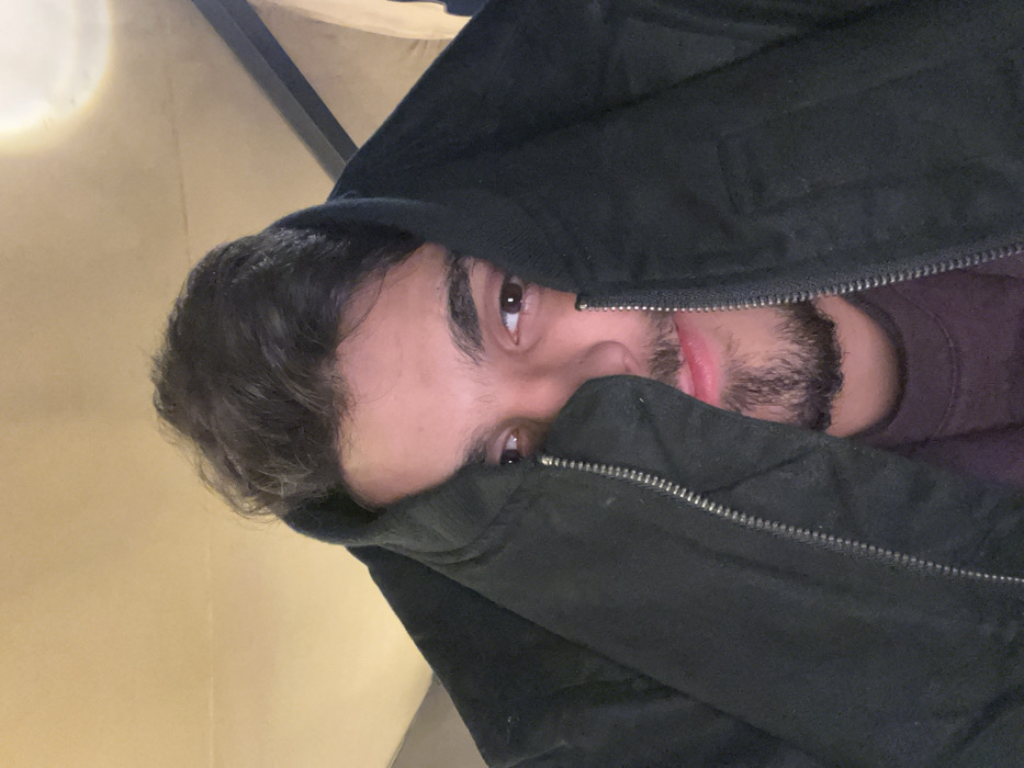 A man with dark hair and a trimmed beard peeks out from beneath the partially zipped-up collar of a black jacket. His eyes, also dark, are visible above the jacket's edge. He seems to be wearing a dark-colored shirt or undershirt. The background appears to be a dimly lit interior with what looks like a light fixture in the upper left corner. The photograph is slightly rotated counterclockwise.