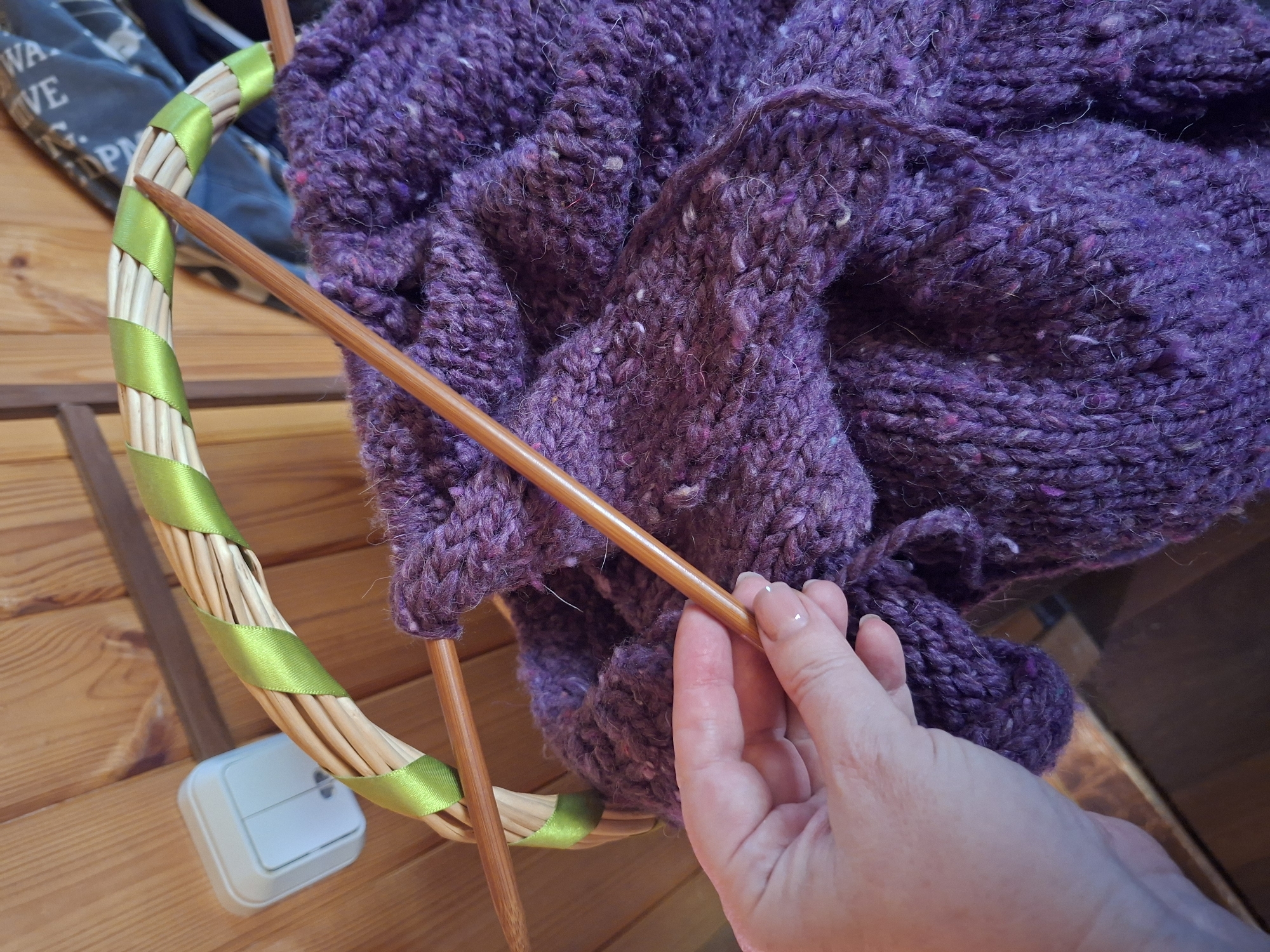 The image captures an overhead view of a person knitting in an indoor setting in Kondratovo, Russia. The central focus is a person's left hand, with light pink nail polish, holding a long, light brown wooden knitting needle. The needle is engaged with a large, chunky piece of deep purple knitted fabric. The yarn has a textured, possibly tweed, appearance with visible flecks of white, pink, and other subtle colors, and a slightly fuzzy quality.
The knitted fabric is draped over and around two wooden knitting needles. To the left of the knitting, a decorative circular object made of light-colored wooden sticks is visible, wrapped spirally with a bright green satin ribbon. Part of the knitted project rests against this circular frame.
The setting appears to be a rustic interior, with a light-colored wooden surface, possibly a table or floor, forming the immediate background. On this surface, a white, rectangular electrical outlet or switch is visible. In the upper left background, behind the circular object, a dark fabric item with partially visible white text can be seen, which reads "WA VE P". Another darker wooden surface is visible in the distant background. The overall scene conveys a cozy and engaged crafting activity.
