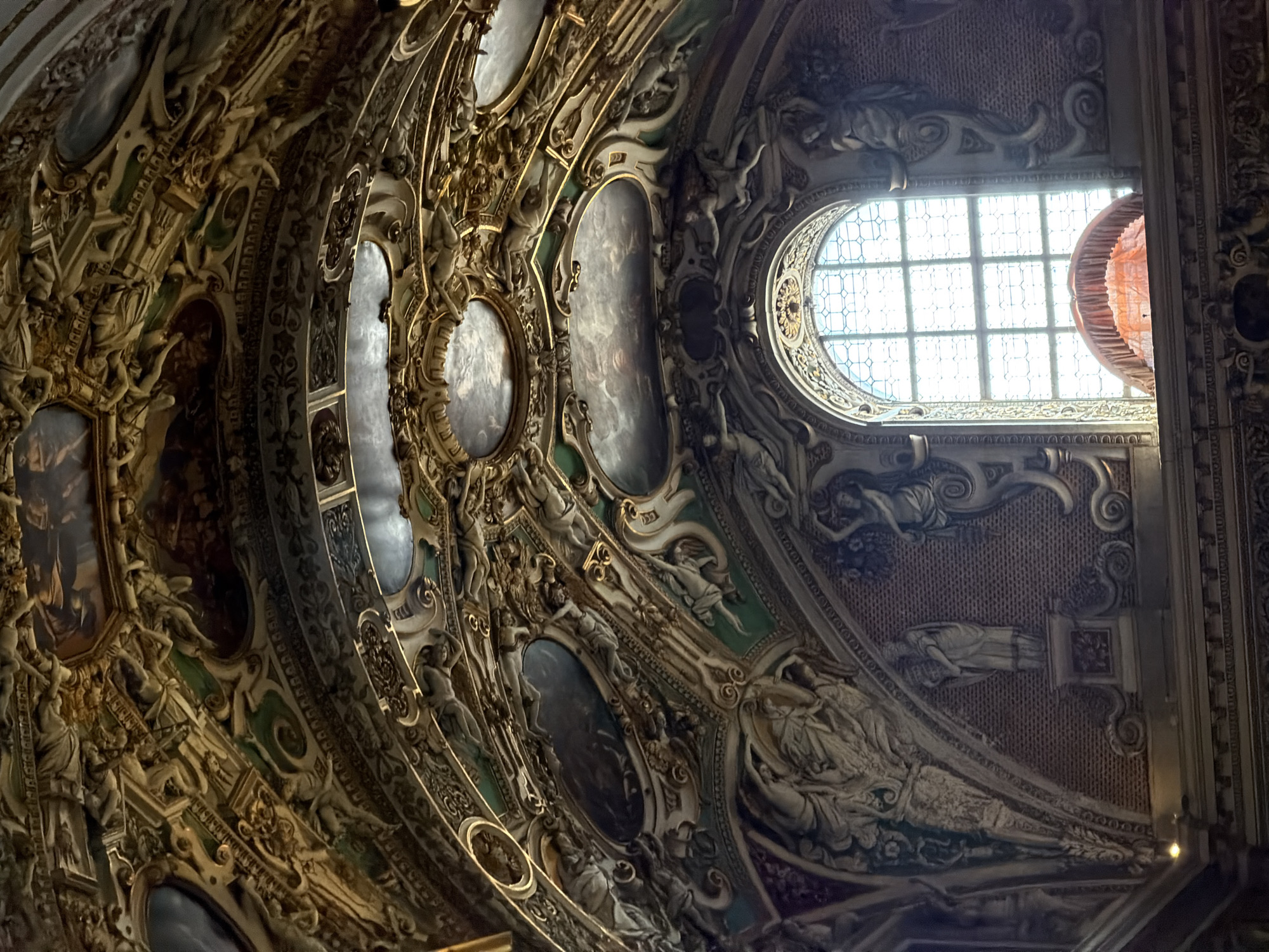 The image shows the interior of a richly decorated ceiling in a building located in Mozzo, Italy. The ceiling is adorned with intricate golden stucco work, featuring various ornamental patterns, scrolls, and sculptural elements. Several oval and rectangular panels depicting paintings of celestial or mythological scenes are embedded within the stucco. The architectural style appears Baroque or Rococo, characterized by its opulence and elaborate ornamentation.
Below the ceiling, a large arched window with a grid-like pattern is visible. The window is brightly lit from the outside, casting a soft glow. In front of the window, an ornate, draped fabric structure, possibly a canopy or curtain, is visible, adding a touch of color with its orange or pink hue. The walls surrounding the window are decorated with relief sculptures of figures, some appearing to be classical statues. The overall impression is one of grandeur and historical significance, indicative of a church, palace, or other important historical building.