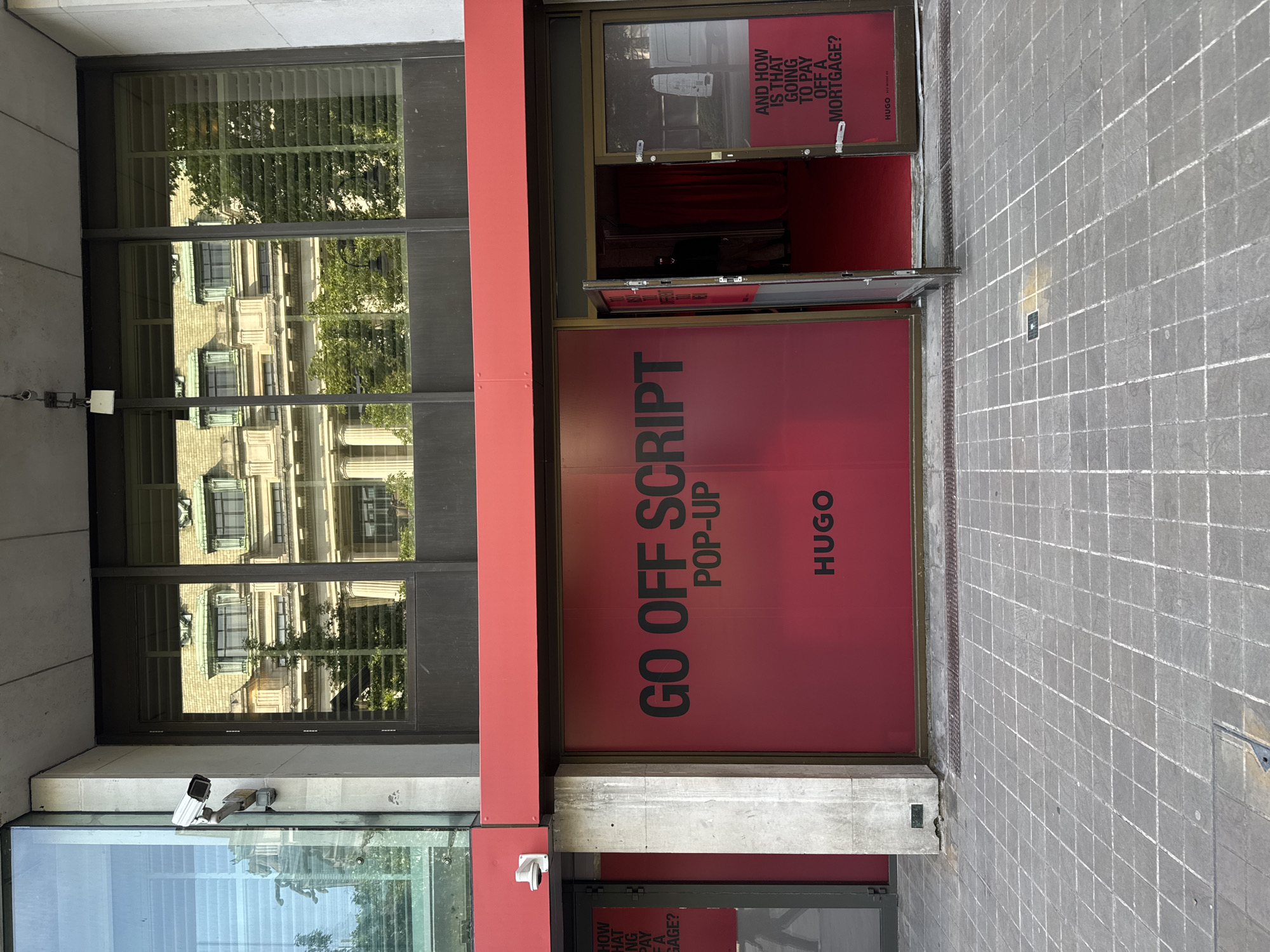 Here is a description of the image.
The image shows the front of a Hugo Boss "Go Off Script" pop-up store located in London, United Kingdom. The store has large red signage with the words "GO OFF SCRIPT POP-UP HUGO" in black letters. The store entrance is partially open, revealing a red interior. On the window is the phrase "AND HOW IS THAT GOING TO PAY OFF A MORTGAGE?". There is a window to the left of the store entrance that reflects the building across the street. A security camera is mounted on the building above the storefront. The sidewalk in front of the store is paved with gray square tiles.