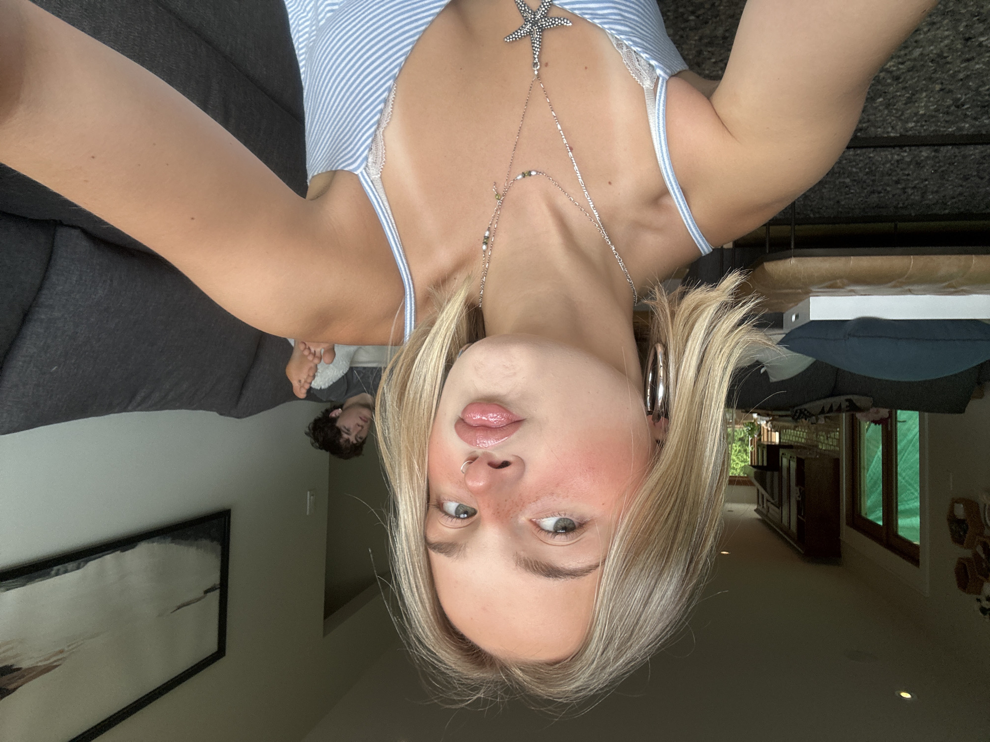 The image depicts a young blonde woman taking a selfie indoors, in what is stated to be a residential location in Squamish, Canada. The woman, positioned in the foreground, faces the camera with a puckered-lip expression and direct eye contact. Her short-to-medium length blonde hair frames her face, and she exhibits noticeable freckles across her nose and cheeks, accentuated by rosy blush. She wears a nose ring on her left nostril and large silver hoop earrings. Her attire consists of a light blue and white striped camisole with visible white lace trim, complemented by layered silver necklaces, one featuring a prominent starfish pendant. Distinct tan lines are visible on her shoulders and upper chest.
In the background, to the right of the woman, a young man with dark, curly hair is partially visible, reclining on a dark grey fabric sofa. He is looking towards the left side of the frame, seemingly observing something off-camera or past the woman. One bare foot rests on a white, textured item, likely a blanket or cushion.
The indoor environment appears to be a living area transitioning into a kitchen. To the left, a doorway with green-tinted glass windows or doors allows a glimpse of outdoor greenery. Further back, dark wood cabinetry and a range hood suggest a kitchen space. To the right, a light grey wall displays a large, rectangular framed abstract artwork with muted tones of white, grey, brown, and dark blue/black. A low, brown leather bench with a dark blue cushion and a white box is situated behind the woman's left shoulder. The floor is covered with a dark, textured material. Natural daylight illuminates the scene, with highlights on the woman's hair and skin.