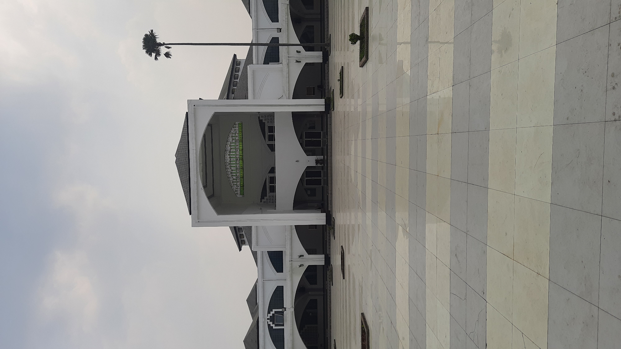 The image shows the exterior of a modern building with a large paved courtyard in front. The building is predominantly white with arched walkways and a prominent central entrance featuring a large archway. The roof is dark and peaked. A tall palm tree stands to the right of the building, silhouetted against an overcast sky. The courtyard is paved with large, alternating light and dark square tiles, some of which appear wet, reflecting the dull sky. Small, square patches of greenery are set into the paving at intervals. Text is visible on the building's facade above some of the archways. This is a view of the Grand Mosque of Bandung, located in Bandung City, Indonesia.