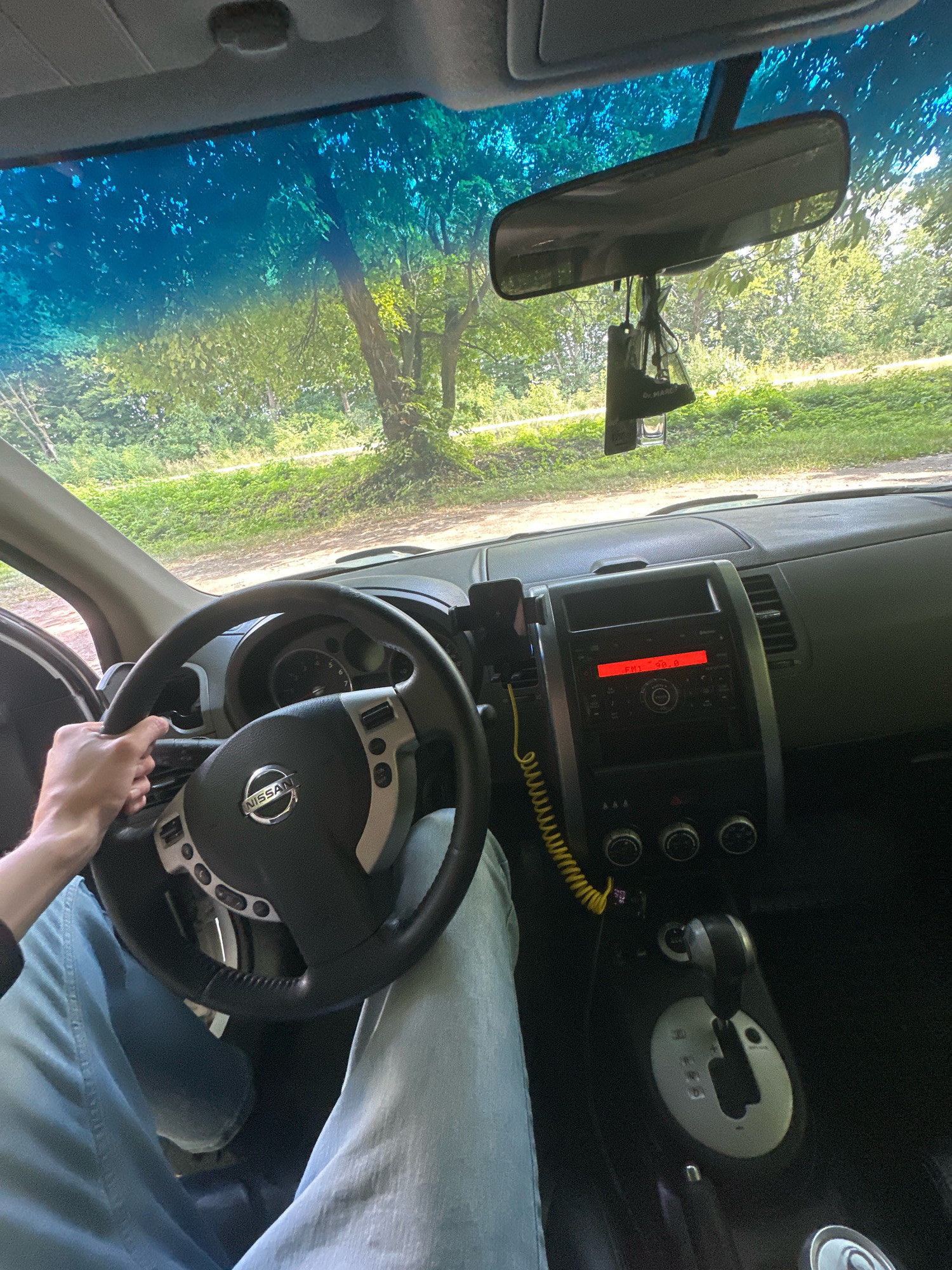 The image shows the interior of a Nissan X-Trail vehicle, viewed from the perspective of the driver. The driver's hands are on the steering wheel, and their legs are visible, wearing light blue jeans. The dashboard features a radio display showing "FM1 98.8", a gear shifter, and climate control knobs. A phone mount is attached to the dashboard. Outside the vehicle, through the windshield, a tree-lined road is visible, suggesting a rural or semi-rural setting. The lighting indicates it is daytime. The location is identified as Shatava, Ukraine.