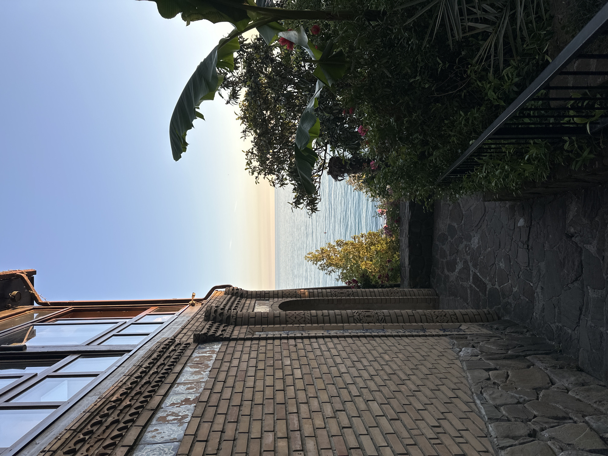 This image depicts a serene coastal landscape in Sochi, Russia, captured from a low vantage point looking upwards. On the left, a multi-story building crafted from light brown bricks occupies a significant portion of the frame. It features large windows with dark frames, reflecting the clear sky. The building's facade is intricately detailed with decorative brick patterns and sections of light-colored ceramic tiles featuring complex designs.
To the right of the building, a dense and lush array of green vegetation creates a natural border. This foliage includes prominent broad, banana-like leaves, various shrubs, and trees adorned with small pink or red flowers. Nestled within this greenery on the far right, a dark metal railing and a stone-paved path or staircase are partially visible, suggesting an elevated walkway.
Beyond the building and vegetation, a calm, blue-green sea extends horizontally, meeting a clear, light blue sky. The sky brightens towards the horizon, indicating either early morning or late afternoon with a soft, warm light. The weather is clear and sunny, contributing to a tranquil and peaceful atmosphere. No specific activity is taking place, and no text is visible in the image.