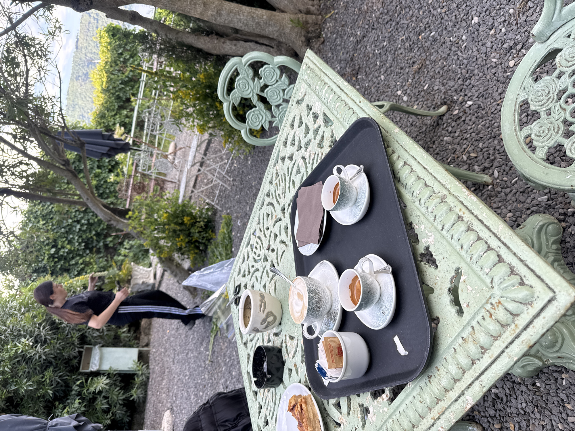 The image presents an outdoor scene in Castel Gandolfo, Italy, focused on a light green, intricately designed metal table and chair set placed on a gray gravel surface. On a black tray sitting atop the table are four cups, two of which appear to contain espresso, one with a cappuccino, and another with sugar packets. There are also saucers, spoons, a napkin, a small vase, an ashtray, and a plate with what appears to be a pastry.
In the background, a person dressed in black sportswear with white stripes is visible, standing near vegetation and a decorative white metal structure that may be part of a garden or terrace. The landscape includes trees and hills, suggesting a scenic overlook, likely a characteristic view of Castel Gandolfo. The lighting suggests a daytime setting.