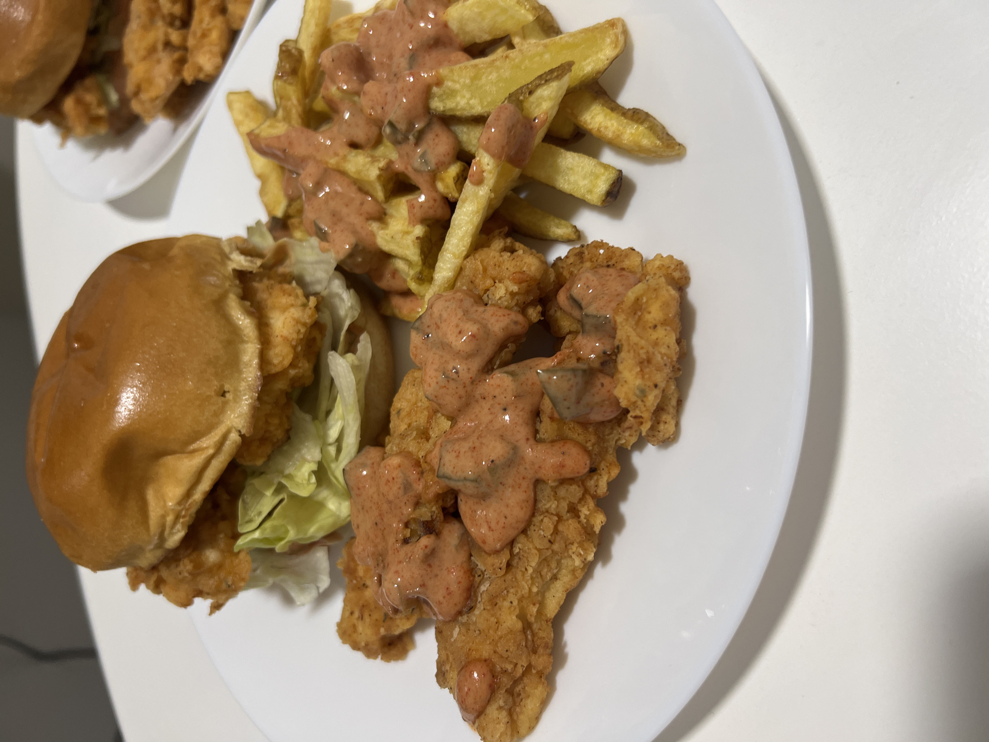A white plate containing a chicken sandwich with lettuce, fries, and a piece of fried chicken is on a white surface. The sandwich has a shiny bun, fried chicken, and lettuce. The fries and chicken are covered with a sauce. A second plate containing fried chicken is partially visible in the upper left corner. The location is not identifiable as Mogán, Spain from the visual information provided.