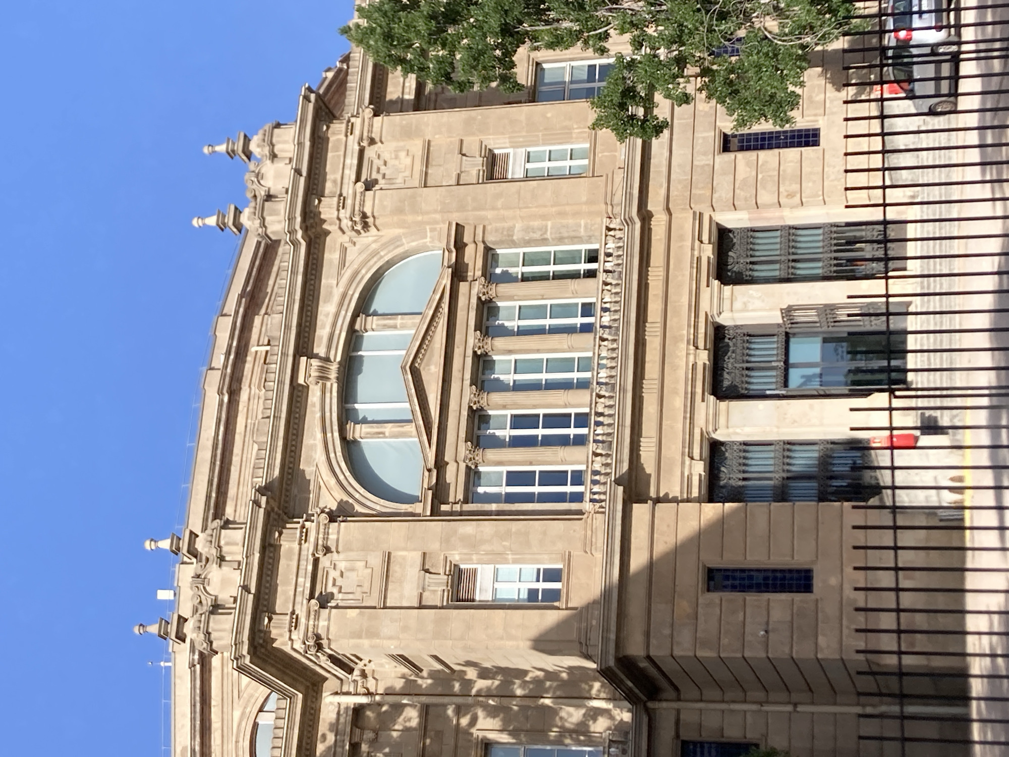 This is an outdoor, eye-level, medium shot of the Palau de la Generalitat de Catalunya. The building, constructed from light brown stone, displays neoclassical architectural elements. The facade features arched windows with multiple panes and pilasters. A balcony with a railing runs across the upper level. The sky is clear and blue. In the foreground, a black metal fence partially obstructs the view of the building. To the right, a lush green tree with full foliage partially obscures the building's right side. Two cars are visible behind the fence, a silver hatchback and a white and silver SUV, both parked. The sun casts shadows on the building.