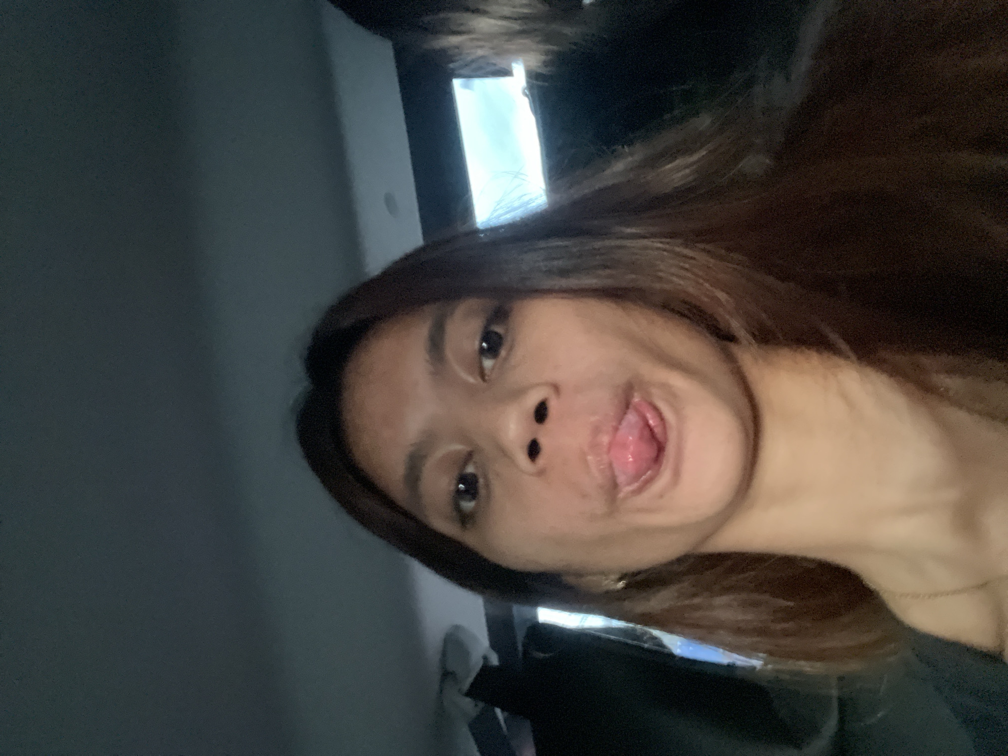 A woman with brown hair is centered in the image, sticking her tongue out towards the camera. She has tan skin and is wearing what appears to be a dark-colored top. A gold chain is visible around her neck. The background includes a blurred interior with a window that lets in light. The location is potentially Davao City, Philippines, based on external context but not discernible from the image itself.
