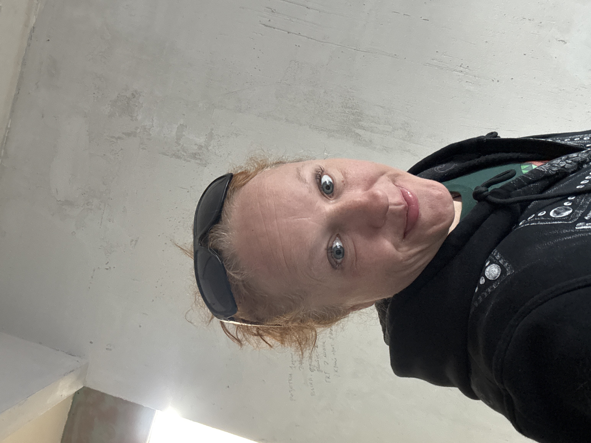 A woman with fair skin and light reddish hair is angled downwards, looking at the camera. She has light blue eyes and is wearing sunglasses pushed up onto her head. She is wearing a dark hooded sweatshirt, possibly with some embellishments visible on the shoulder. The background appears to be an unfinished or undecorated interior wall, likely in a building. There appears to be some writing on the wall. The location is Chaikovsky, Russia. The scene does not offer any other details.