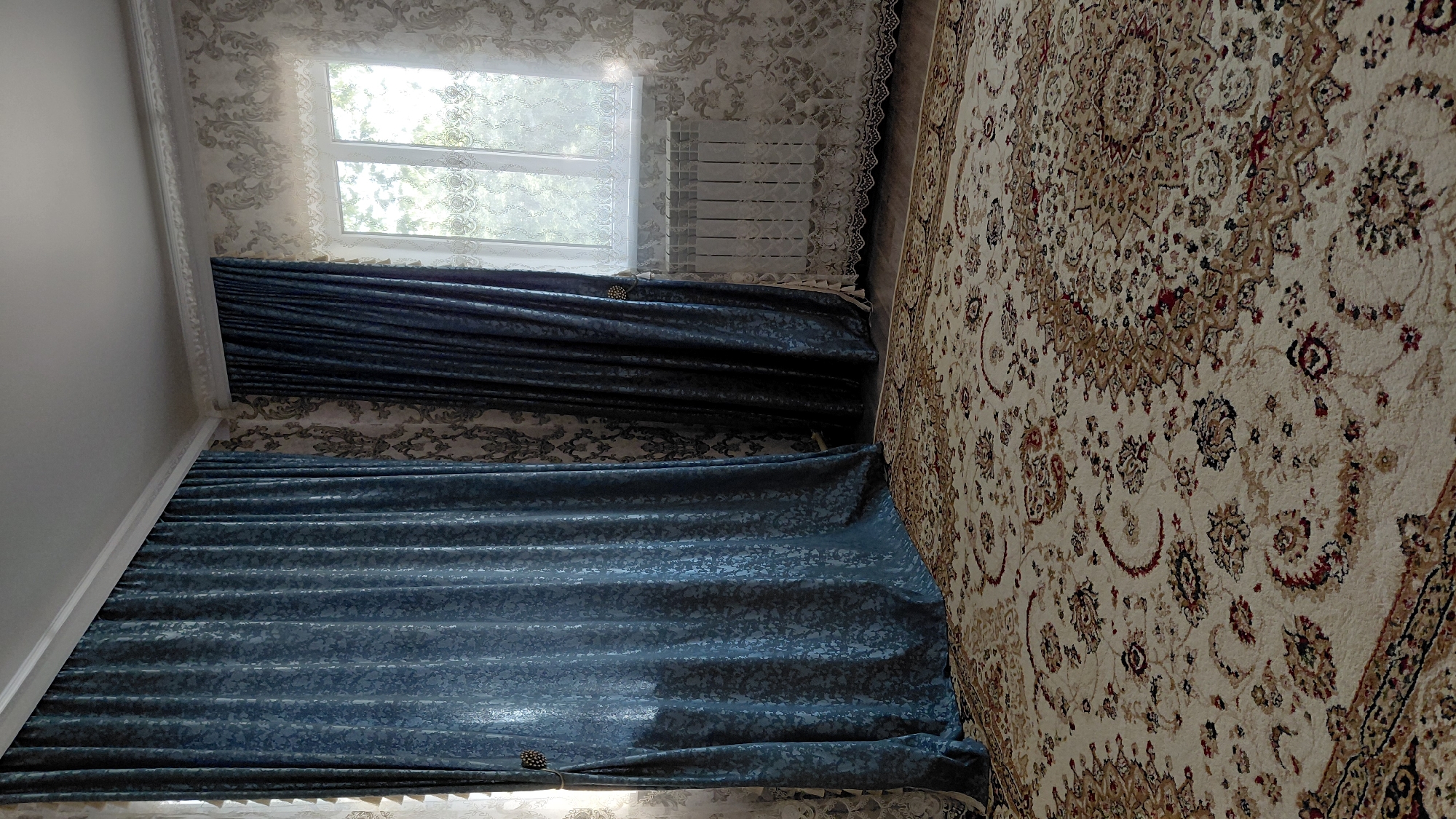 The image shows a corner of a room with a large, ornate rug on the floor. Two sets of heavy, blue curtains hang from the ceiling on the left side of the image, one set partially obscuring a window. The walls are covered in a patterned wallpaper. A radiator is visible beneath a lace-covered window on the right. The room appears to be located in Aktobe, Kazakhstan.