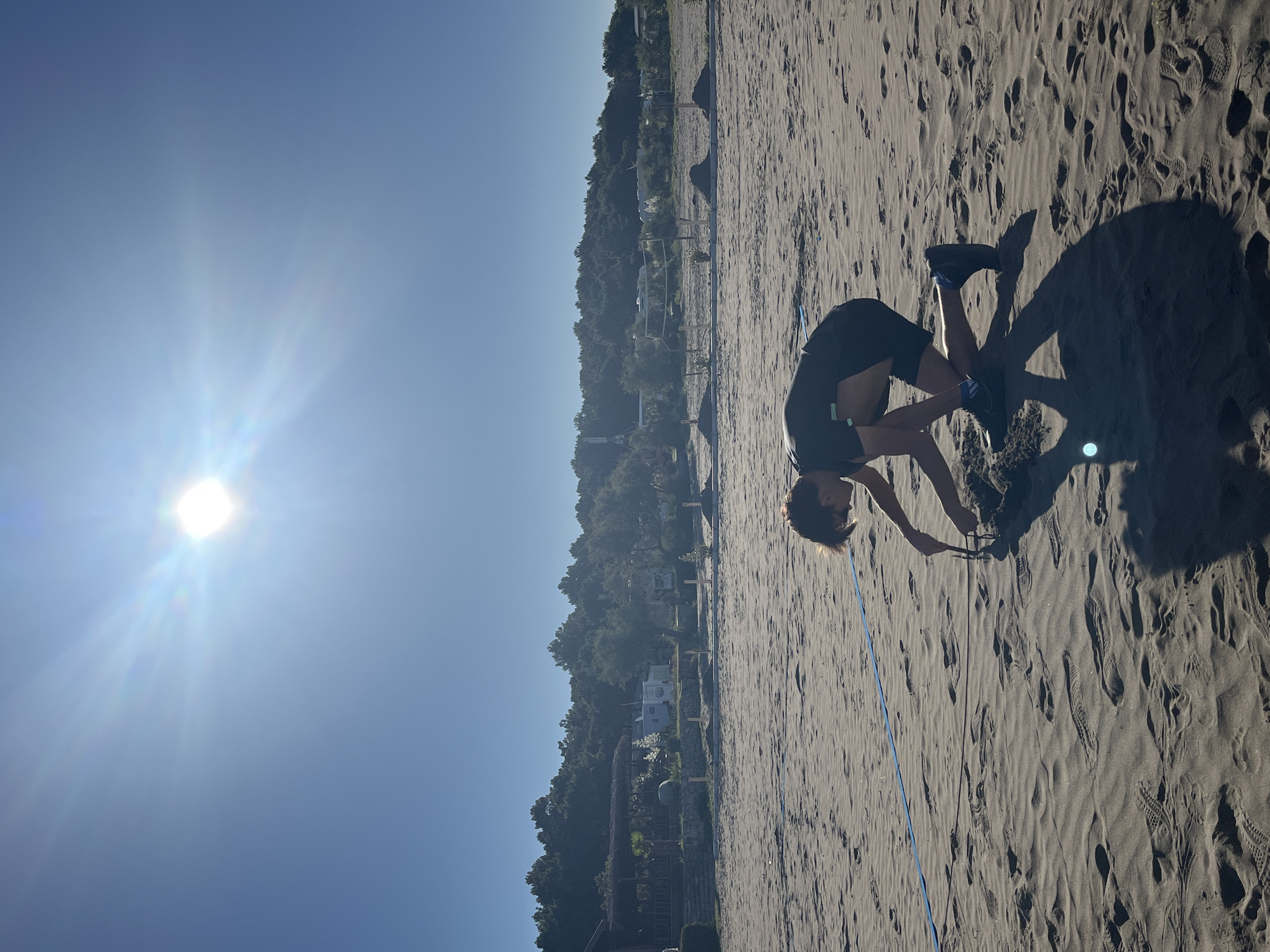 A person is kneeling on a sandy beach, drawing in the sand with a stick. The sun is bright in the sky, and there are trees in the background. The person is wearing a dark t-shirt and shorts. There is a blue line on the sand, possibly marking a boundary or a game. The beach appears to be in a coastal area, with vegetation and some structures visible in the distance. The overall impression is one of a leisurely activity on a sunny day at the beach.