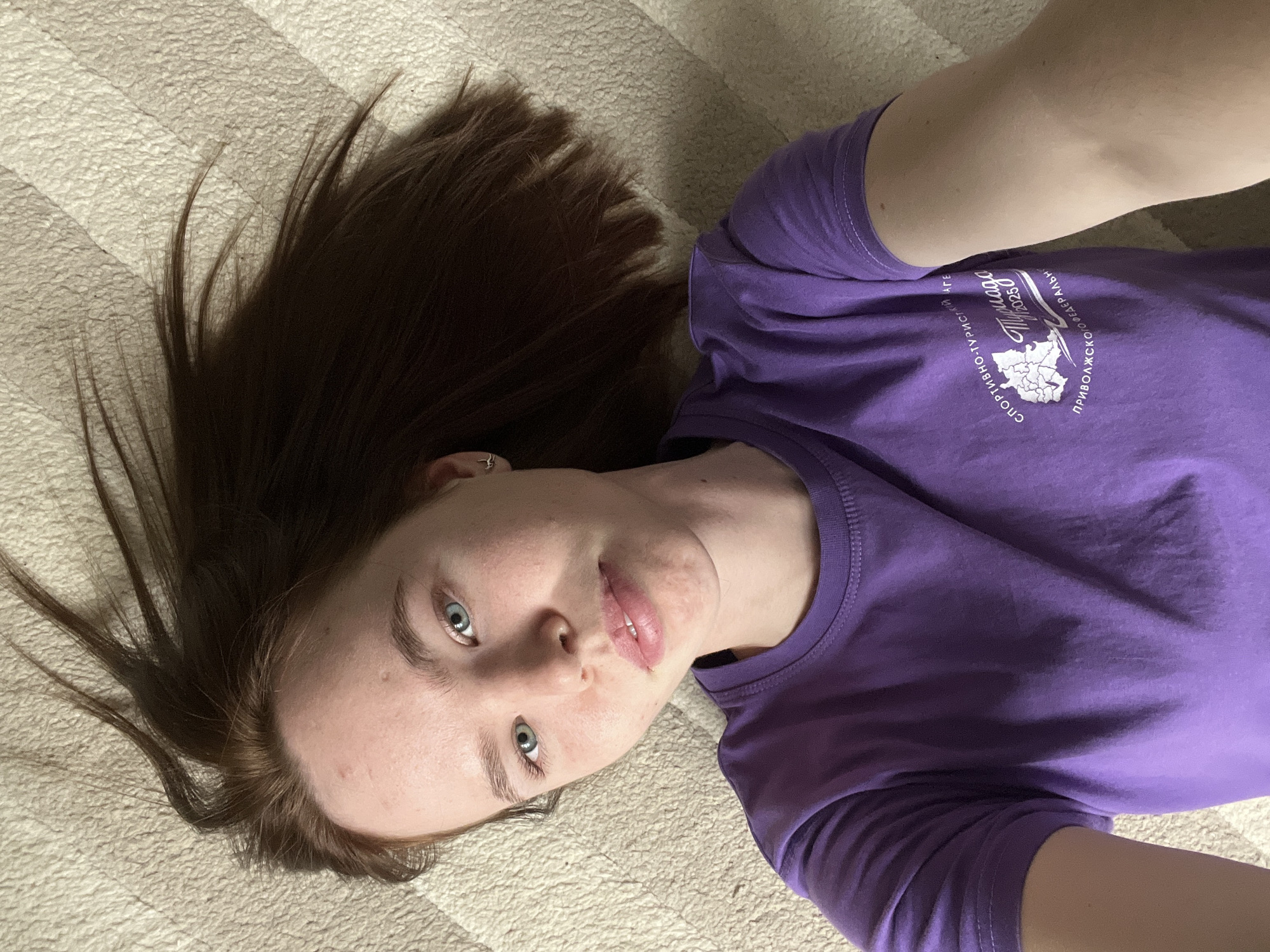 A young woman with long, dark brown hair and blue eyes is captured in a selfie, lying on a light-colored, textured surface that resembles a blanket or rug. Her hair is spread out around her head. She wears a plain purple short-sleeved t-shirt with a white graphic print on the front. The graphic features a stylized map, the year "2025", and Cyrillic text. The visible text reads "ПРИВОЛЖСКОГО ФЕДЕРАЛЬНОГО" (Volga Federal District), "Тулуга" (Tuluga), and "СПОРТИВНО-ТУРИСТИЧЕСКИЙ ЛАГЕРЬ" (Sports and Tourist Camp), indicating a "Tuluga Sports and Tourist Camp of the Volga Federal District 2025". She looks directly at the camera with a neutral, calm expression, her lips slightly parted. A small ring piercing is visible in her left ear. The setting is indoors, with ambient lighting. The provided context for the location is Chaikovsky, Russia.