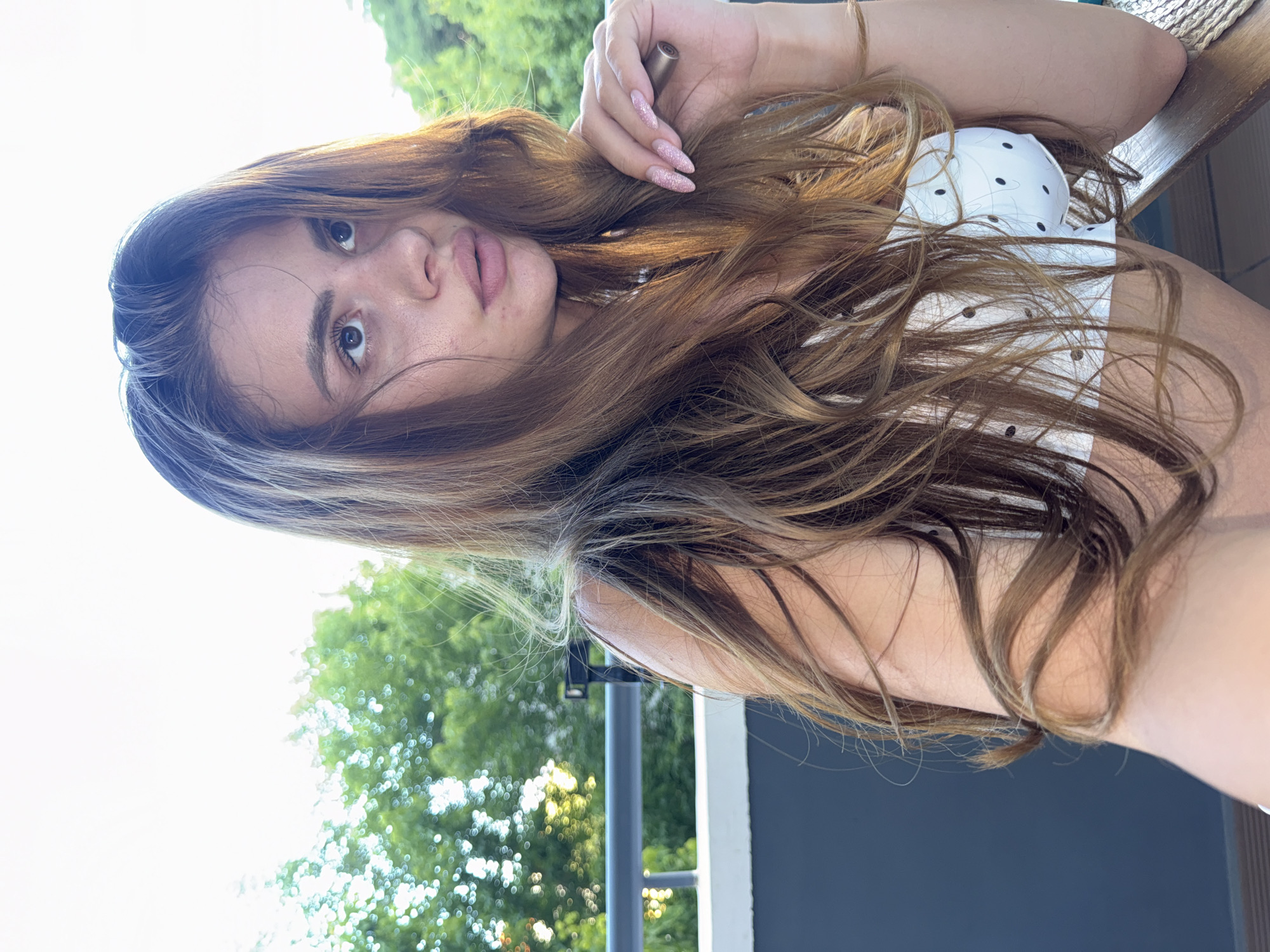 A young woman with long, light brown hair is pictured from the chest up, seated outdoors. She is wearing a white top with black polka dots and has light pink nail polish. Her gaze is directed to her right, away from the camera. In her right hand, she holds a small, metallic object. Behind her, a railing and lush green trees are visible, suggesting an outdoor balcony or patio setting. The lighting indicates it is daytime, with the sun casting a warm glow. The background appears to be residential, with trees suggesting a park or garden area. This scene is consistent with a location in Plovdiv, Bulgaria, given the typical residential architecture and greenery often found in its neighborhoods.
