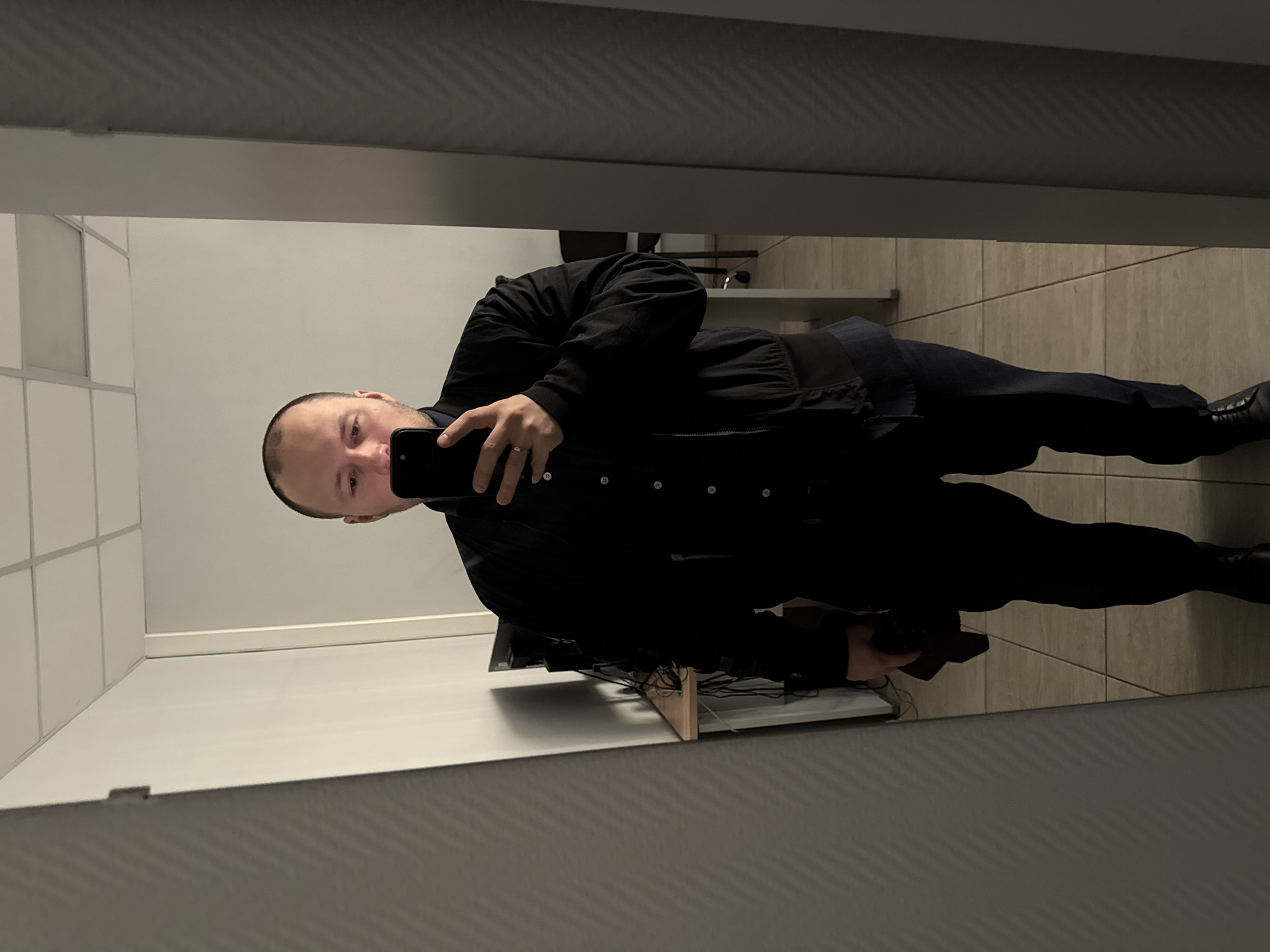 A man is taking a selfie in a mirror. He is wearing a black jacket and pants, and black shoes. He is holding a black smartphone. The background is a tiled wall and floor. There is a desk with cables and a chair in the background. There is no evidence to determine the location is Krasnoyarsk, Russia.