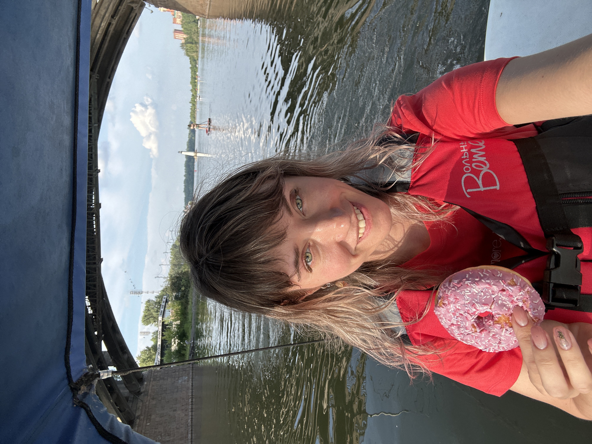 A woman with blonde hair and green eyes is taking a selfie on a boat under a bridge. She is wearing a red shirt and a life vest. In her left hand, she holds a pink donut with white sprinkles. The water is calm, and there are other boats and people on paddleboards in the background. The bridge is made of concrete and spans across the river. The sky is cloudy with patches of blue. The location is unknown.