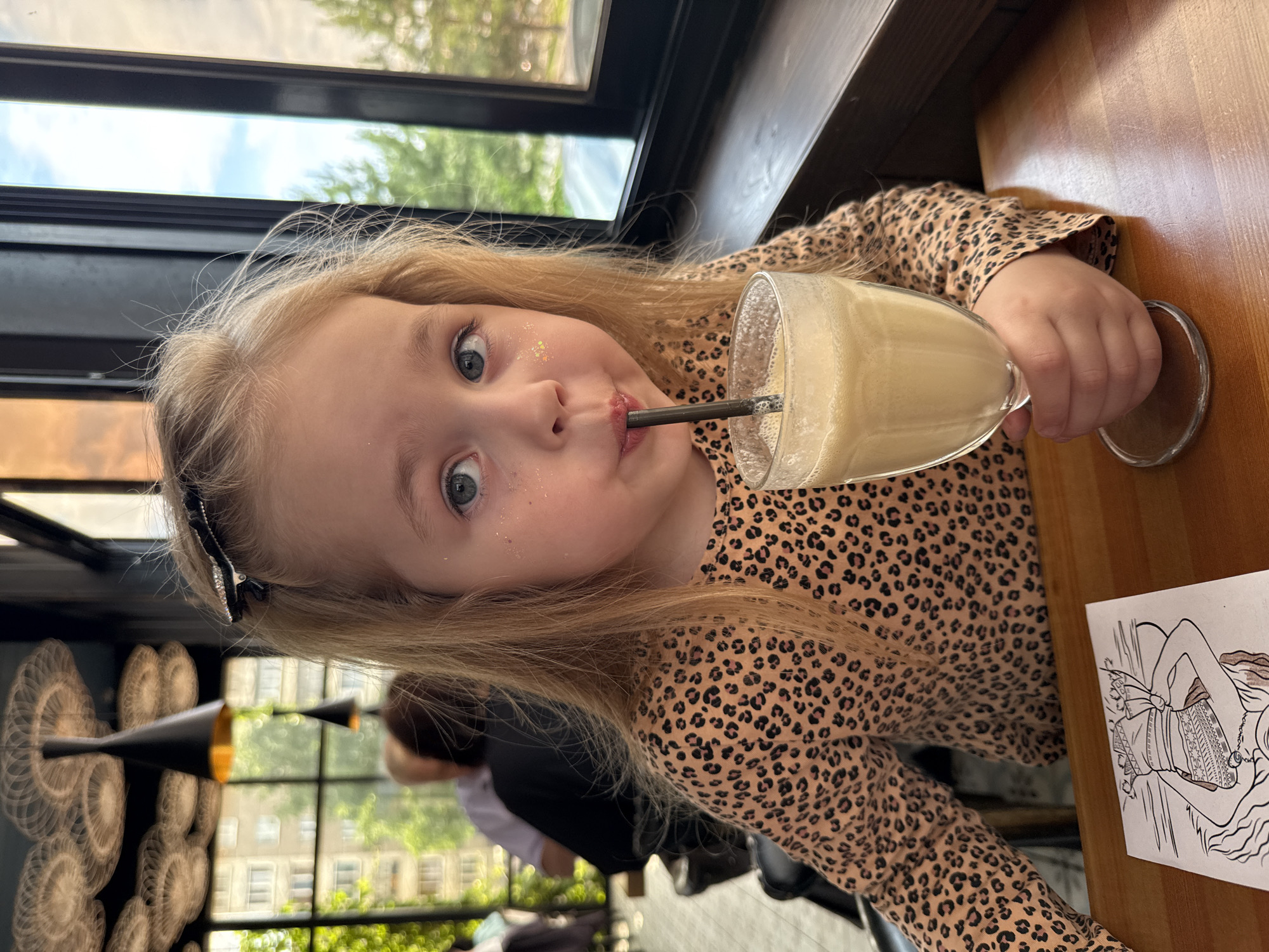 A young girl with blonde hair and blue eyes is drinking a milkshake through a straw from a glass. She is wearing a leopard-print top and has glitter on her face. She is seated at a wooden table in what appears to be a cafe or restaurant. In the background, there are other patrons and decorative lamps. A coloring page is visible on the table in front of her. The scene suggests a casual outing, possibly in Izhevsk, Russia, given the presence of buildings and greenery visible through the windows.