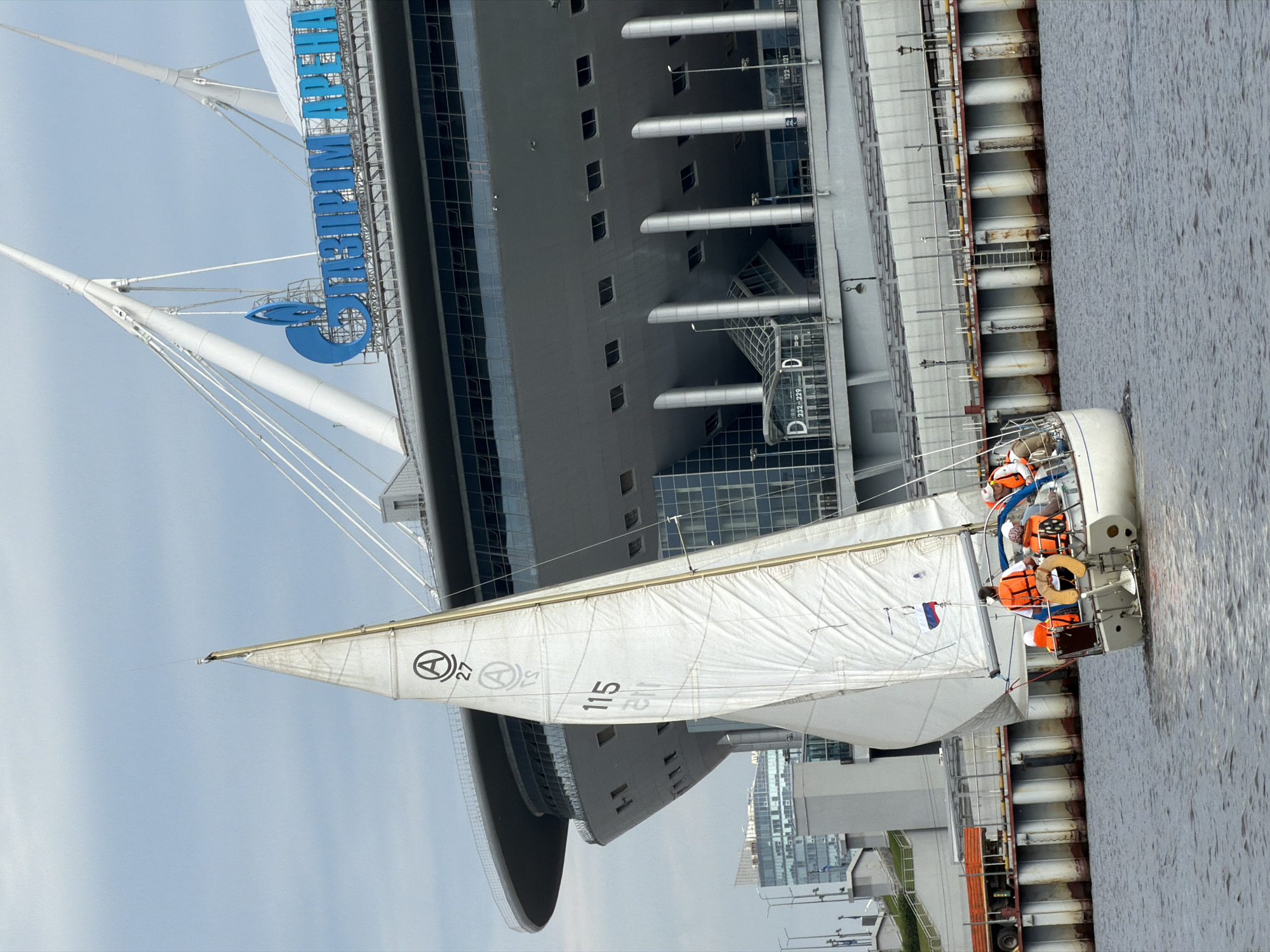 The image depicts a white sailboat with several people aboard sailing on a body of water in Saint Petersburg, Russia. The sailboat has a large white sail with markings including "A 27" and "115". On the boat, people are wearing orange life vests. In the background, a large modern stadium structure is visible, identified by the prominent blue text "ГАЗПРОМ АРЕНА" (Gazprom Arena). The stadium has a distinctive architectural design with white support structures. The sky is overcast with clouds. A pier with decorative fencing and vertical elements runs along the waterfront in front of the stadium. Other buildings, including a modern high-rise, are visible further in the distance. The overall scene is set in an urban waterfront environment.
