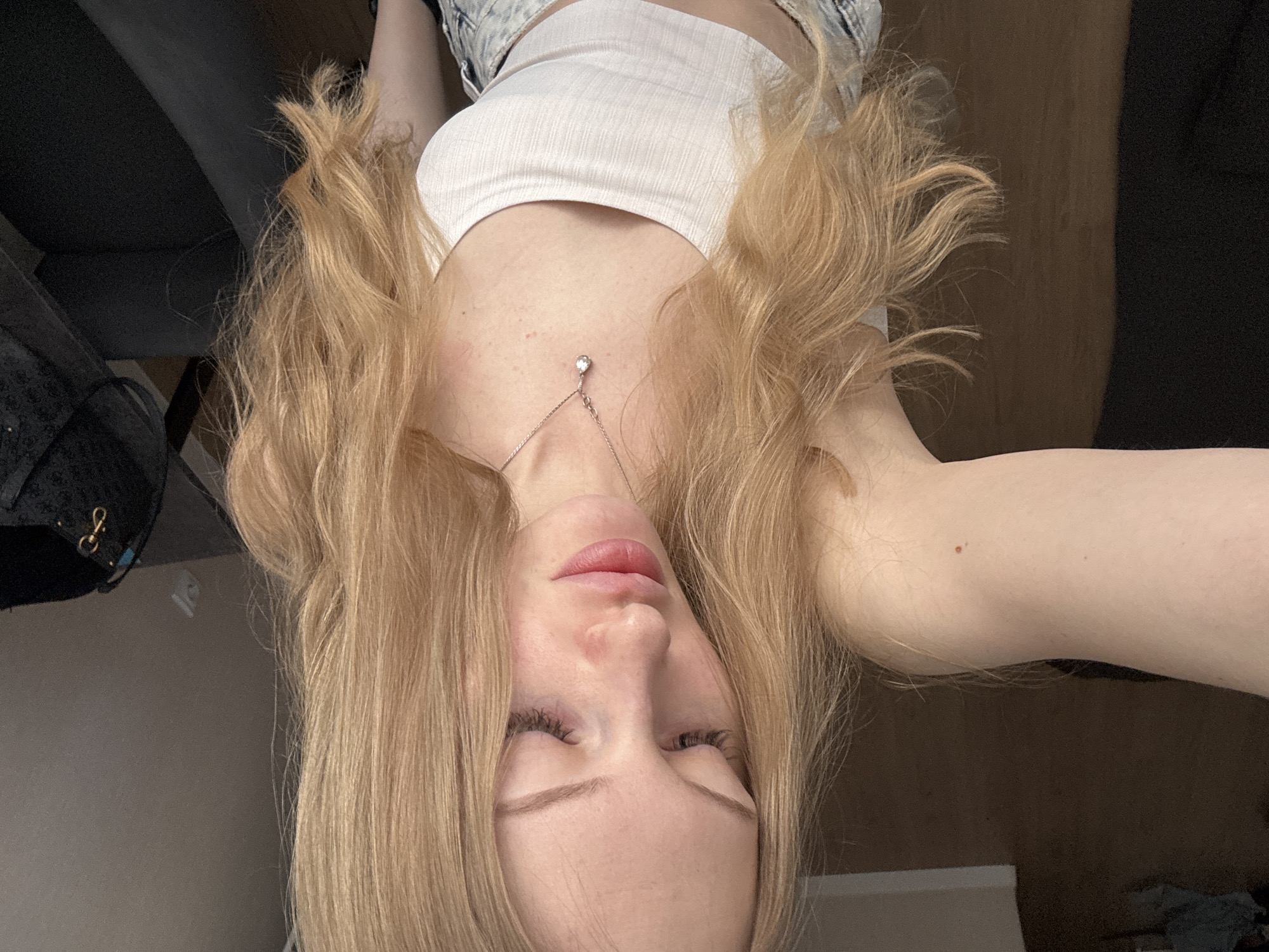 A woman is shown in a selfie-style shot, looking downward with her eyes partially closed. She has long, light blonde hair framing her face and is wearing a white ribbed tank top and a delicate silver necklace with a pendant. The background is blurry and indistinct, showing a mix of dark and light surfaces, including what appears to be furniture. There is no indication that this photo was taken in Perm, Russia.