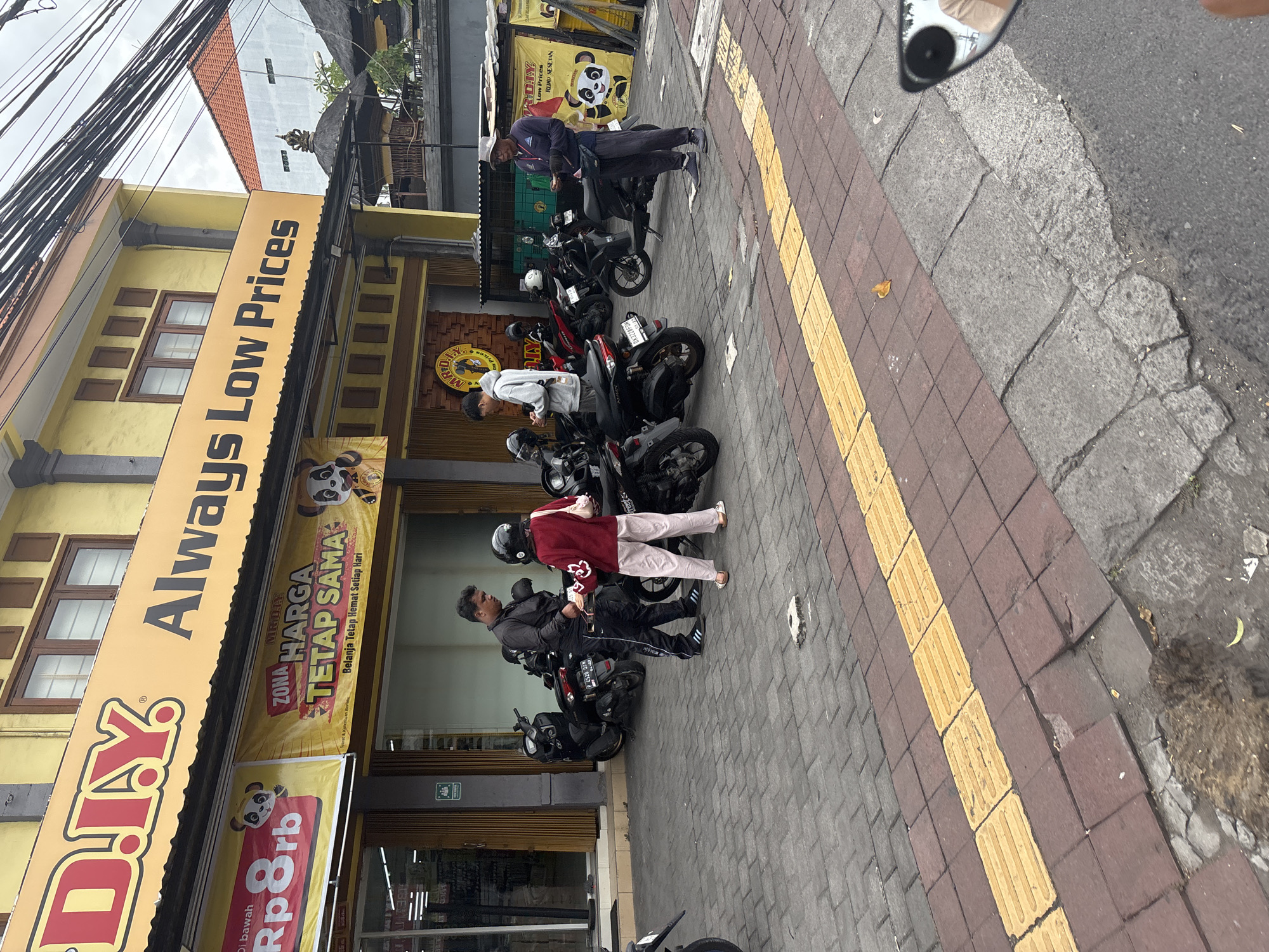 In Denpasar, Indonesia, on a paved sidewalk in front of a yellow building with a large yellow sign that reads "D.I.Y. Always Low Prices," a group of people are gathered around several parked motorcycles. The building has two stories, with windows on the upper level and a storefront at ground level. A banner on the building also advertises "ZONA HARGA TETAP SAMA" (Zone of the same price) and features a cartoon panda character.
There are at least four people visible. One man in a black jacket and black pants with white stripes stands near a woman in a maroon jacket and light-colored pants. They appear to be interacting. Another man in a light-colored hoodie is looking at his phone. A third man in a blue long-sleeved shirt and dark pants, wearing a hat, is also visible, holding something in his hands.
Several black motorcycles are parked on the paved area. The sidewalk has red brick pavers and a tactile paving strip of yellow square tiles. In the background, there are more buildings, including one with a traditional thatched roof, and power lines crisscross overhead. The scene depicts a typical street view in a commercial area.