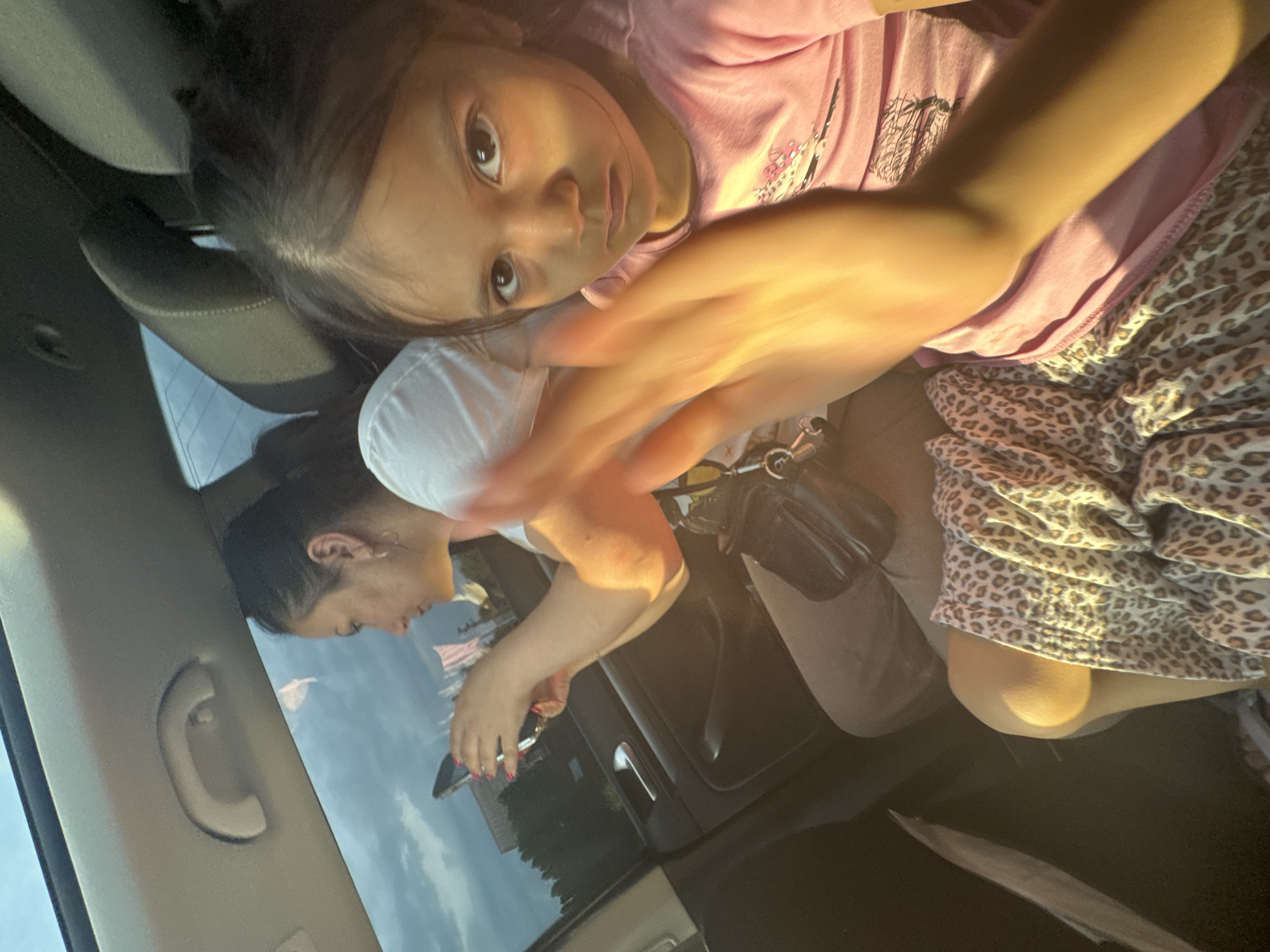 An interior shot captures two individuals inside a moving vehicle, illuminated by strong, warm sunlight suggesting late afternoon or early evening. In the immediate foreground on the right, a young child, likely female, with dark hair and eyes, gazes directly at the camera. She wears a pink t-shirt with a faint graphic design. Her right hand is raised and blurred by motion, partially obscuring her face, and is strongly backlit by the sun. A leopard print blanket or garment is draped across her lap. In the midground on the left, an adult female with dark hair pulled back is visible in profile. She wears a white t-shirt and holds a smartphone horizontally with both hands, appearing to interact with it while looking out the window. Her fingernails are painted pink. A dark shoulder bag or fanny pack rests near her lap. The vehicle's interior features a light-colored headliner with a grab handle, dark door panels with a chrome handle, and dark seats. Outside the window, a blue sky with some clouds is visible above distant green foliage and indistinct vertical structures, possibly flags or banners. The specific location in Suprunivka, Ukraine, cannot be determined from the visual information present in the image.