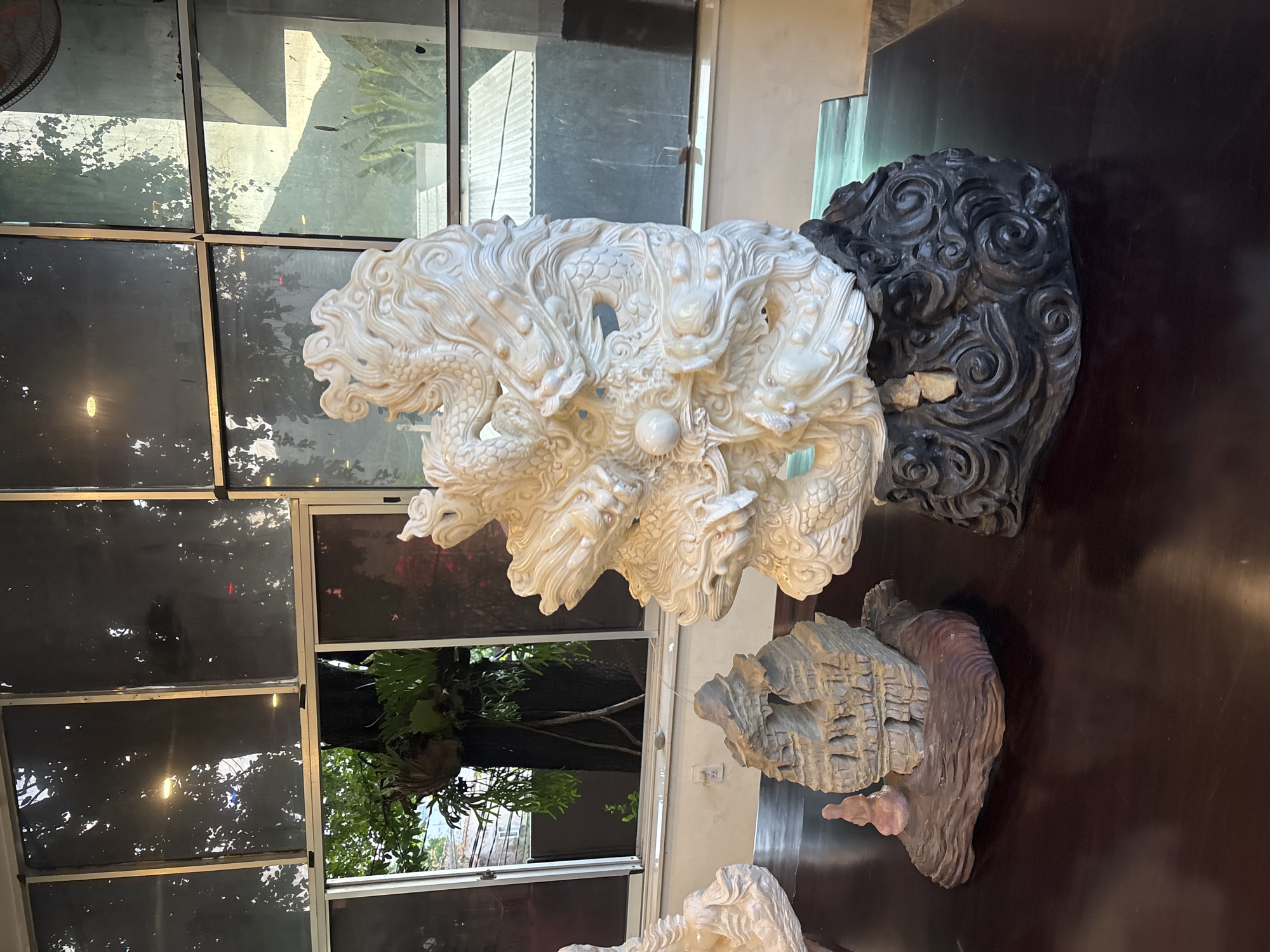 A close-up, vertical shot displays intricate sculptures on a dark, polished surface, likely a table. In the foreground, a large, white sculpture features multiple coiled dragons, their scales and serpentine bodies meticulously carved. A luminous orb sits nestled among the dragons. Behind this, a dark, heavily textured sculpture resembles a swirling vortex or a stylized head. Further back, and to the right, a lighter, layered sculpture evokes rocky formations or ancient architecture. The scene is lit from above, casting subtle shadows and highlighting the detailed textures of the artwork. Reflections of lush greenery and abstract shapes, possibly from windows or other surrounding elements, are visible on the dark surface, suggesting an indoor setting with natural light. The overall mood is one of artistic display, showcasing detailed craftsmanship.