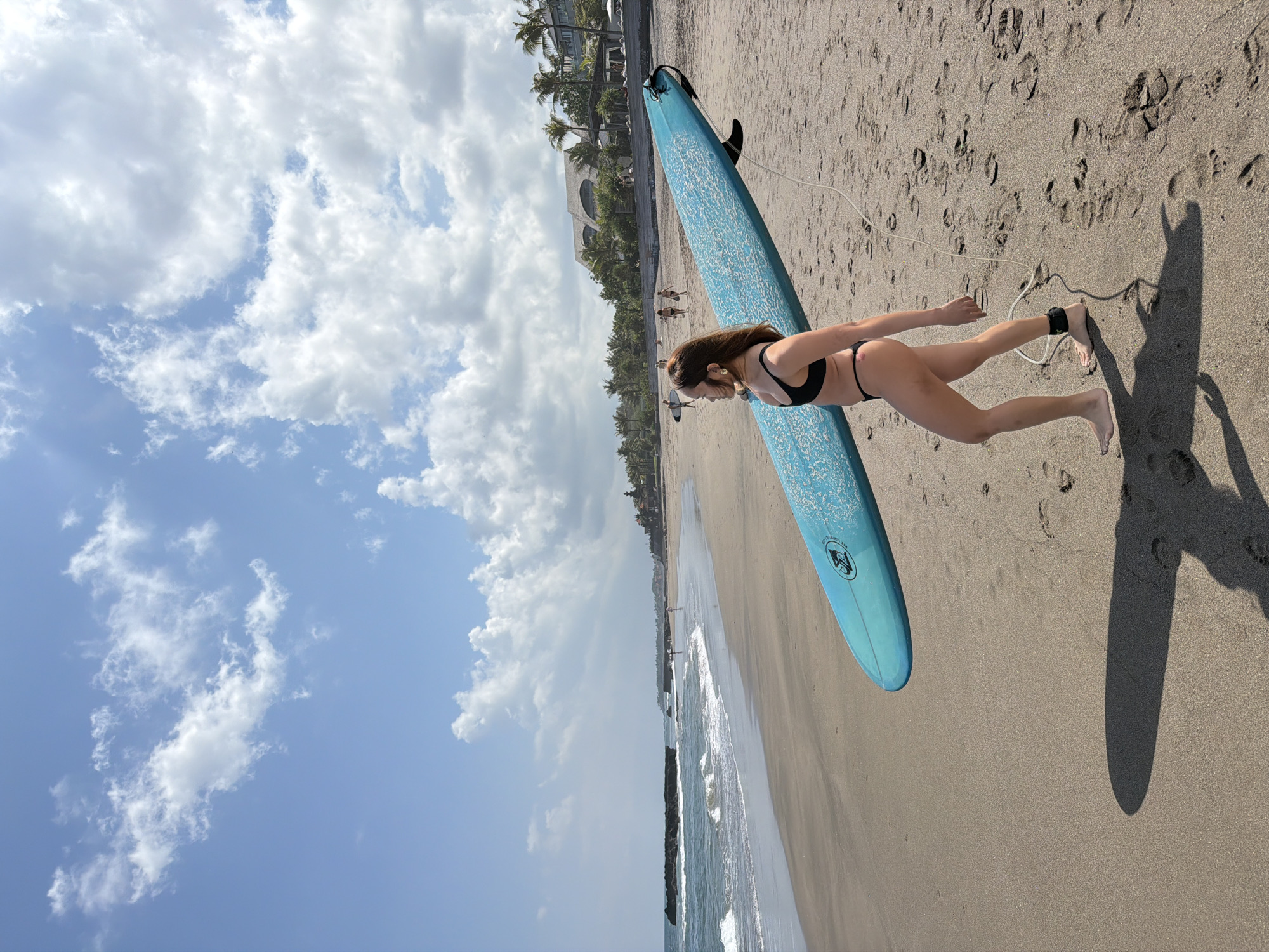 A woman walks along the sandy beach of North Kuta, Indonesia, carrying a bright blue surfboard. The woman is wearing a black bikini and has a black ankle strap connected to the surfboard's leash. Her shadow is cast on the sand. The ocean is to her left with waves rolling onto the shore. In the background, palm trees and buildings can be seen. The sky is a clear blue with scattered white clouds.