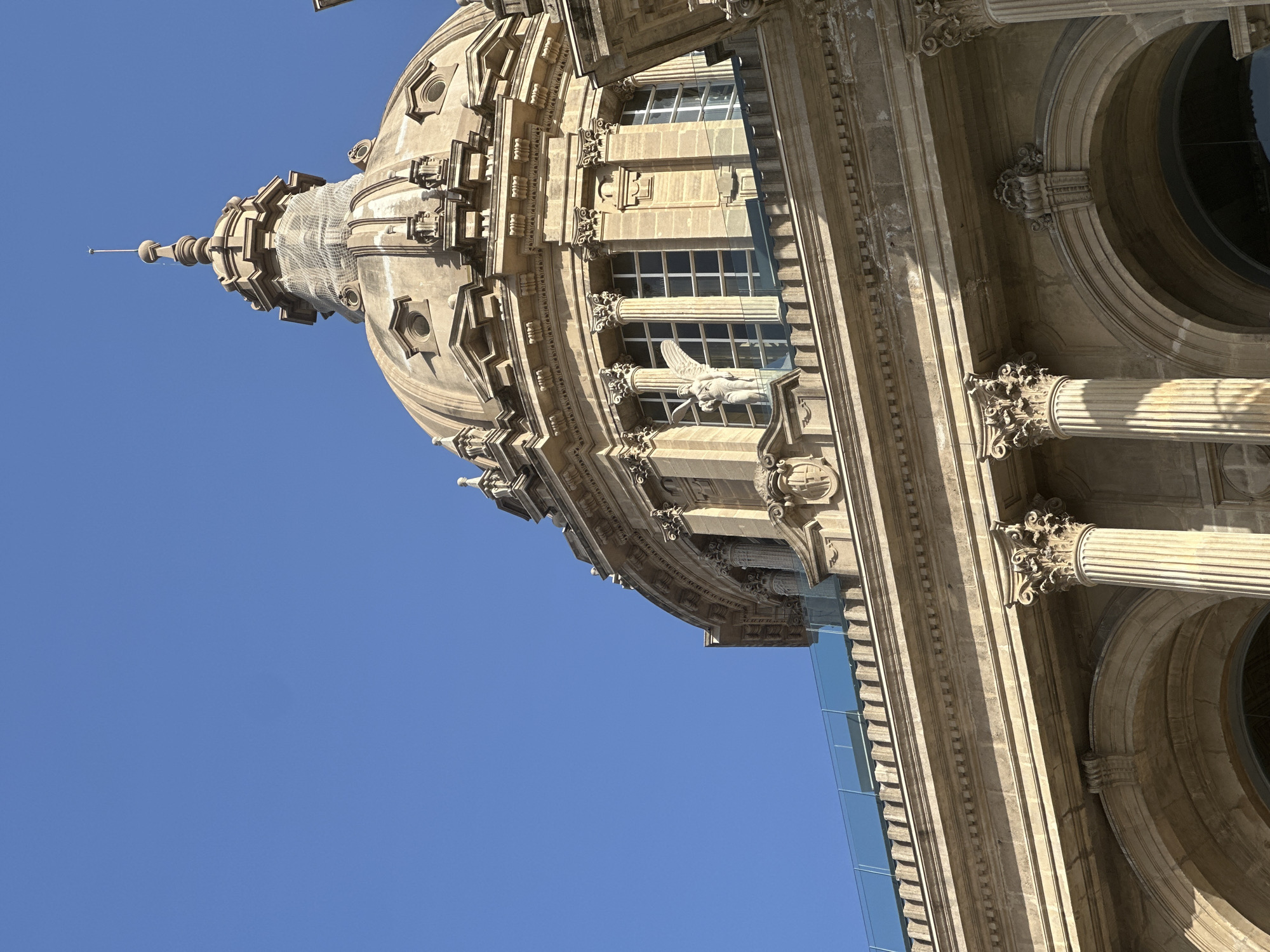 The image shows a close-up of the dome and upper facade of the National Art Museum of Catalonia (Museu Nacional d'Art de Catalunya) located on Montjuïc hill in Barcelona, Spain. The building's stone architecture features classical elements such as columns, arches, and decorative carvings. A prominent dome with a spire is visible against a clear blue sky. A statue of a winged figure stands on a balcony area below the dome, between large windows. The surrounding architectural details are intricate, with carved cornices and pilasters. A modern glass balustrade runs along the top of a lower section of the building. The lighting suggests it is a bright, sunny day.