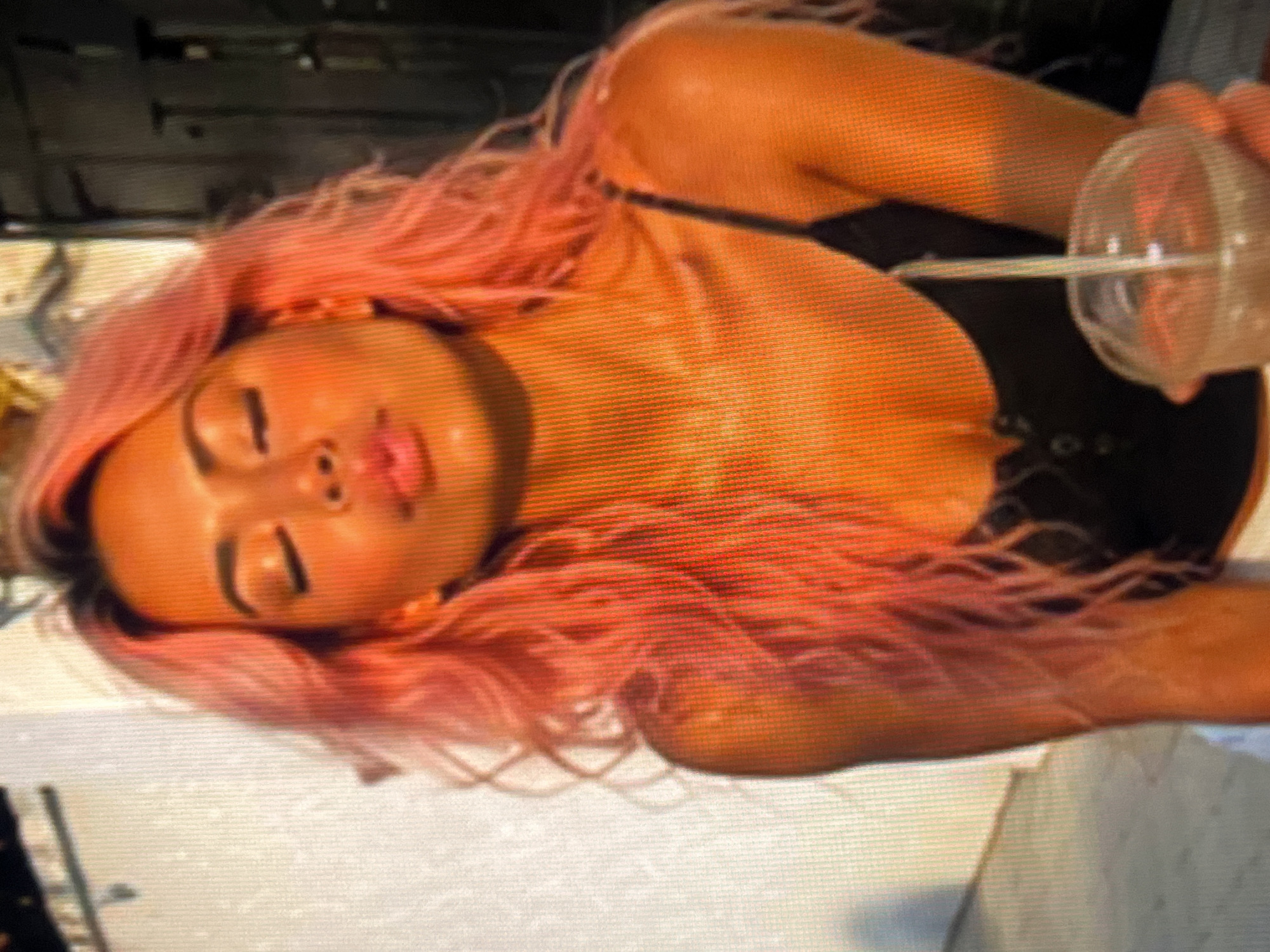 A woman with bright pink hair and closed eyes is captured in a portrait, her face angled slightly upwards with her lips pursed. She is wearing a black tank top. The lighting suggests it is either sunrise or sunset, casting a warm, golden glow on her skin and hair. In the lower right corner of the image, a clear plastic cup with a straw is visible, held by the woman's hand. The background is blurred, with a textured white wall on the left and a darker, out-of-focus area on the right, suggesting an outdoor urban setting.