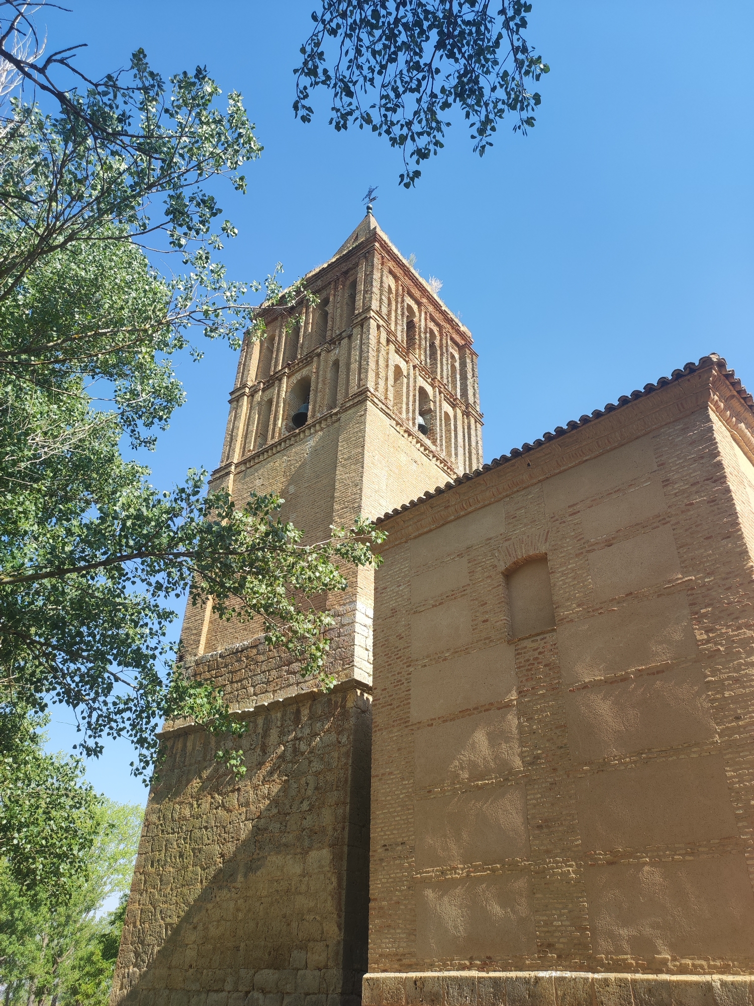 The image displays a view of the church of San Miguel in Abarca de Campos, Spain, under a clear blue sky. In the foreground, lush green trees with visible branches and leaves partially obscure the view of the church tower and its adjacent building. The church tower, constructed primarily of brick, features multiple arched openings and a cross at its apex. The building next to the tower is also made of brick, with a tiled roof. The lower section of the tower and a portion of the adjacent wall are built with stone blocks. Sunlight casts shadows across the stone and brick surfaces. No people or animals are visible in the frame.