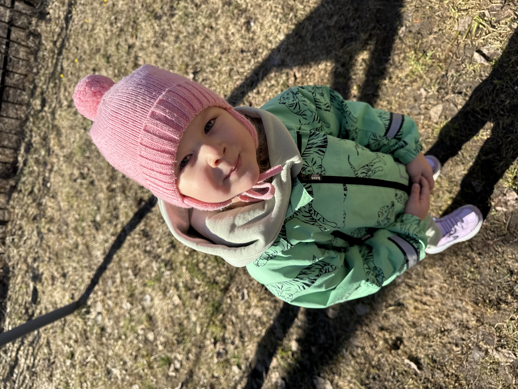 A small child wearing a pink knit hat with a pom-pom, a light green hooded sweatshirt, and a green jumpsuit with black tiger stripes is sitting on the ground in a grassy area. The child is looking up at the camera. The ground is covered in dry grass and leaves. There are some shadows visible on the ground. The location is Ферма, Russia.