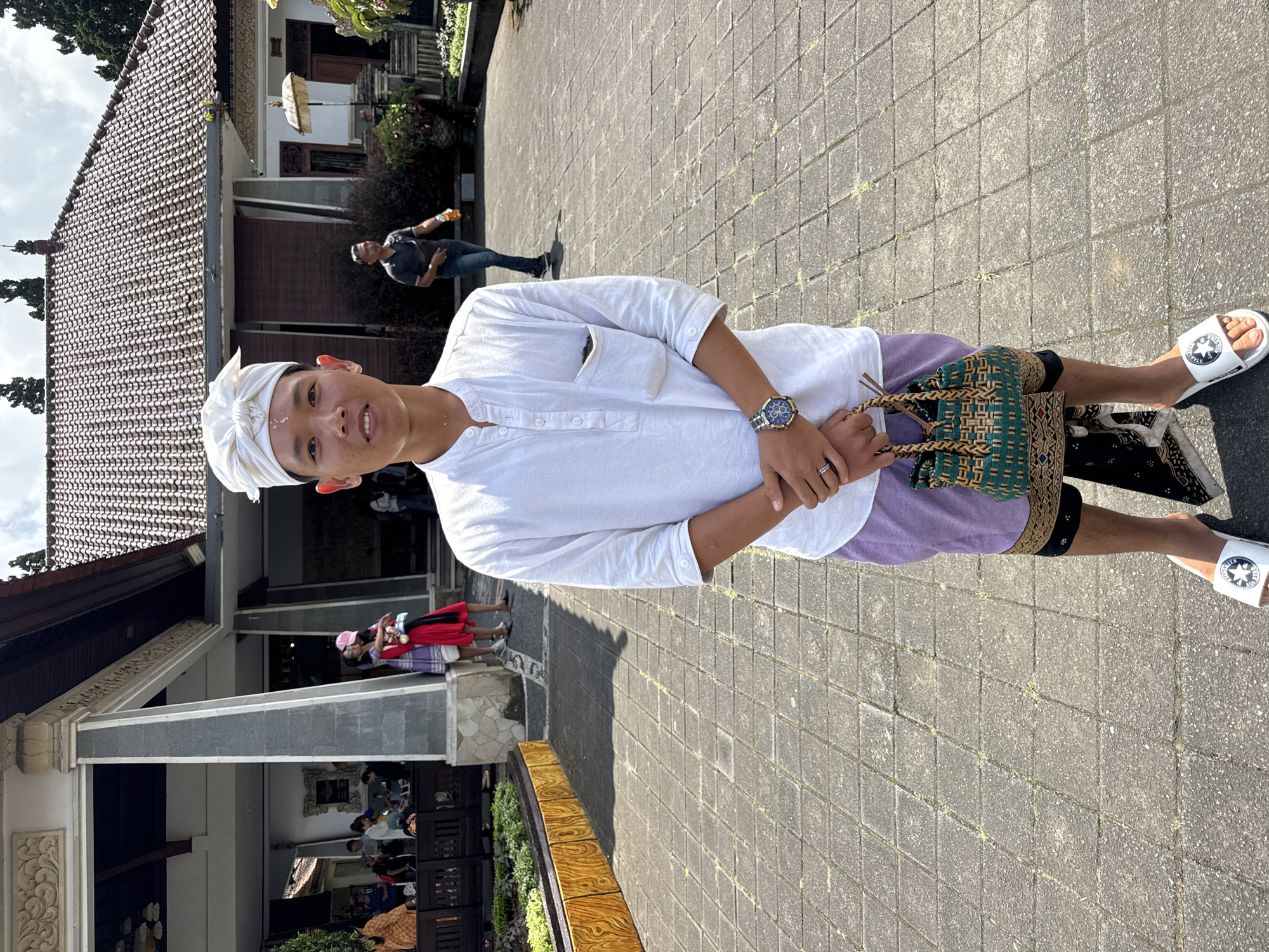 The image shows a person standing on a paved surface in Baturiti, Indonesia. The person is wearing a white shirt, light purple shorts, a white head wrap, and white sandals. They are holding a green and brown woven bag. Buildings with traditional Balinese architecture are visible in the background, along with other people. The scene appears to be outdoors during daylight hours.