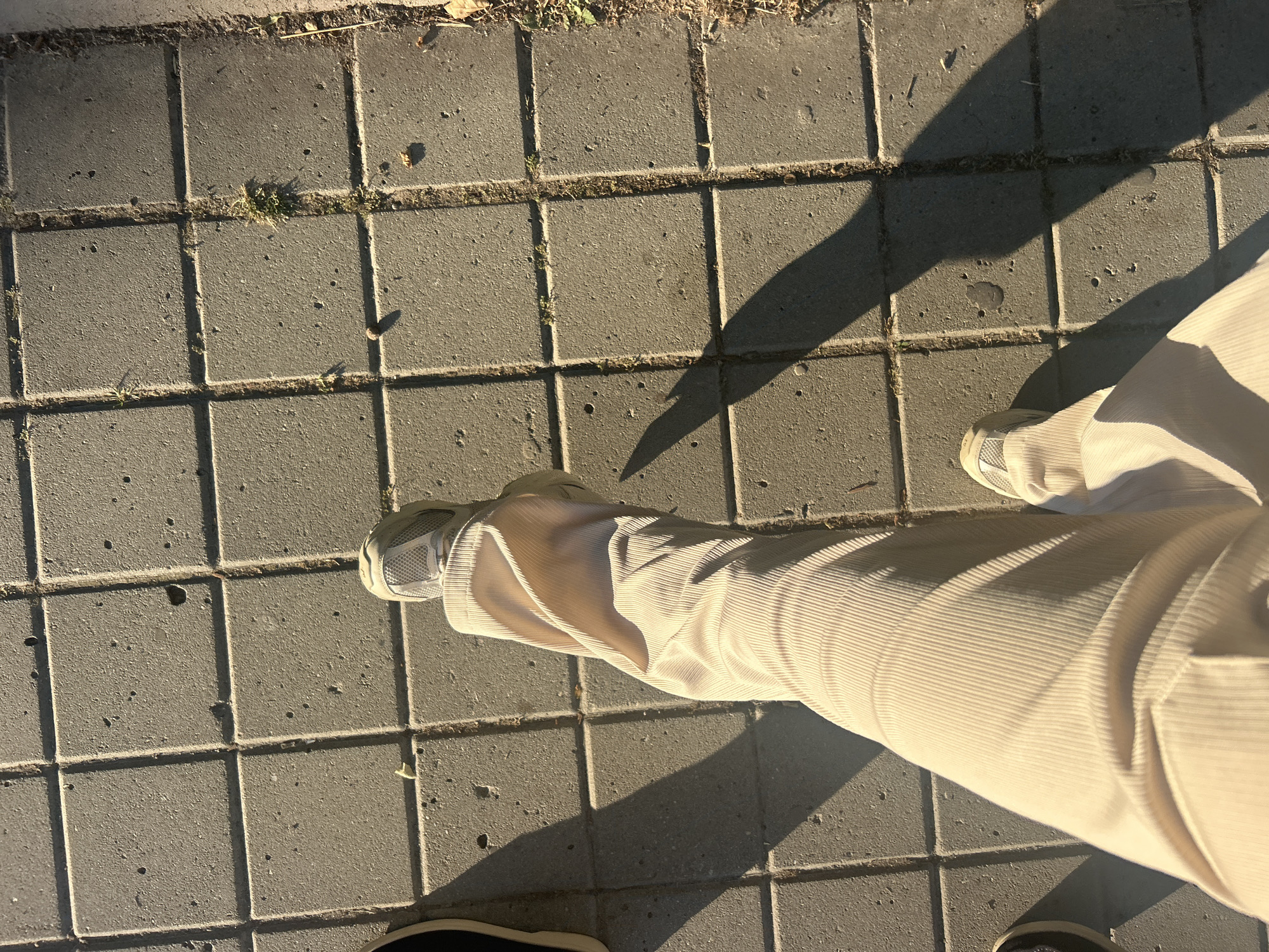 The image shows a person's legs and feet as they walk on a sidewalk paved with square grey concrete tiles. The person is wearing light-colored corduroy pants and white sneakers. A shadow of the person is cast on the sidewalk to their left, indicating the sun is to their right. There are some sparse weeds and debris visible in the gaps between the tiles. The scene is outdoors, with the lighting suggesting it is daytime. The location is described as Unknown, Unknown.