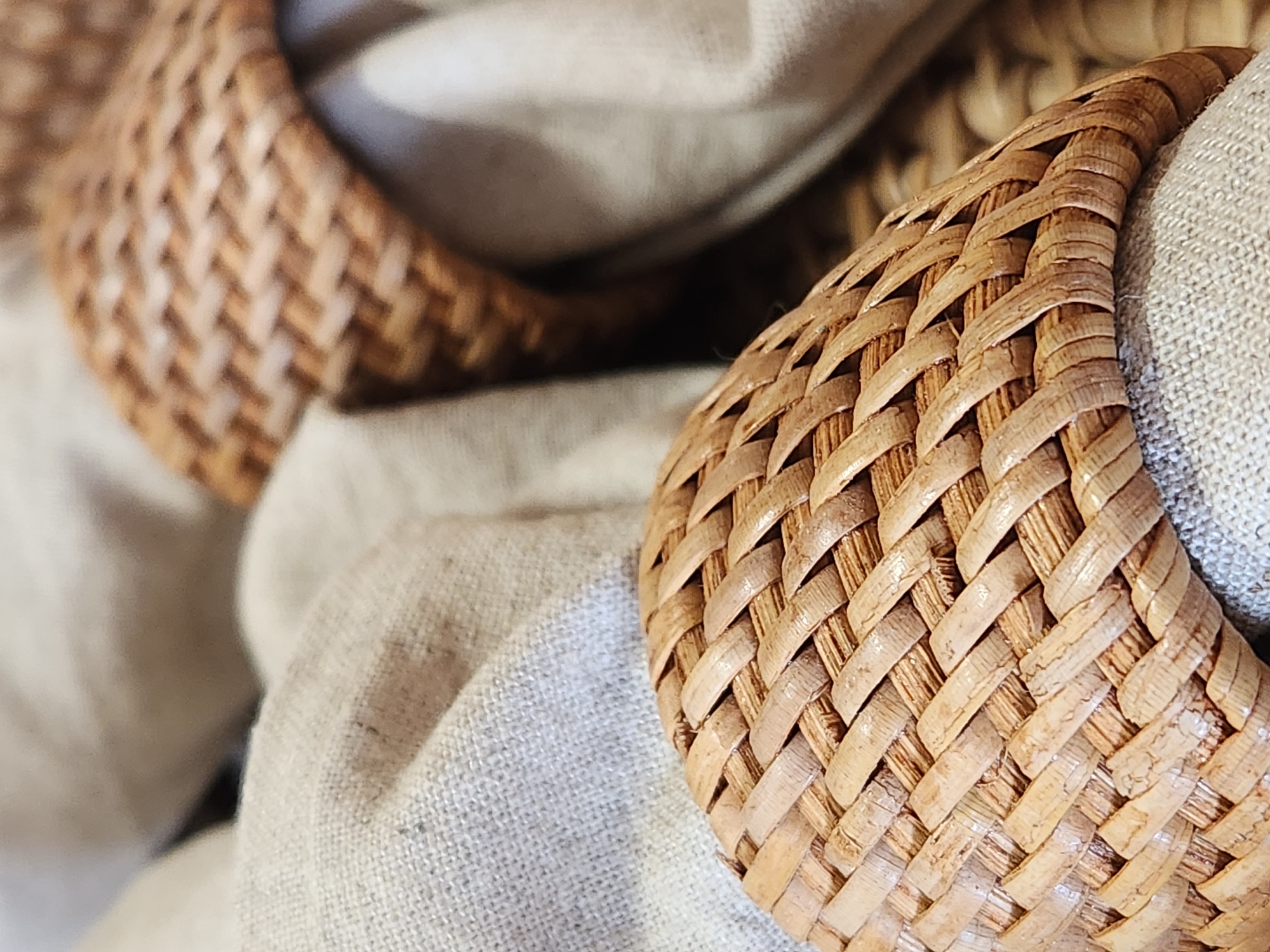The image is a close-up, overhead shot of two woven, light brown napkin rings. The napkin rings are made of rattan or a similar natural fiber, woven in a crisscross pattern. The front napkin ring is in focus and partially obscures the second napkin ring behind it. Both napkin rings appear to be holding light beige or cream-colored cloth napkins. The fabric of the napkins has a visible texture, suggesting it is linen or a similar material. The background is softly blurred, but appears to be more of the same fabric or a woven surface. The lighting is soft and natural, creating subtle shadows that highlight the texture of the rattan. There are no people or any indication of a city or location.
