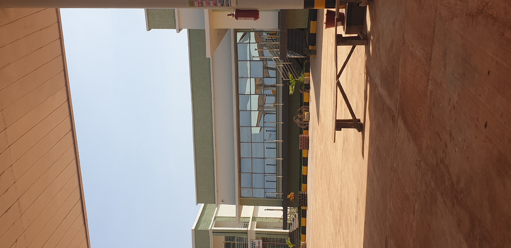 The image captures a daytime outdoor scene in front of a modern building with a green and white facade in Sabon Tasha, Nigeria. The building features a large expanse of glass windows reflecting the sky and parts of the interior structure. To the left, a sign reads "DRINKS DEPOT." A yellow and black striped border runs along the base of the building. In the foreground, a wooden bench is placed on a reddish-brown dirt or concrete surface. Scattered around the area are a few potted plants, some decorative brickwork, and a fire extinguisher mounted on a pole to the right. The sky is clear and blue.