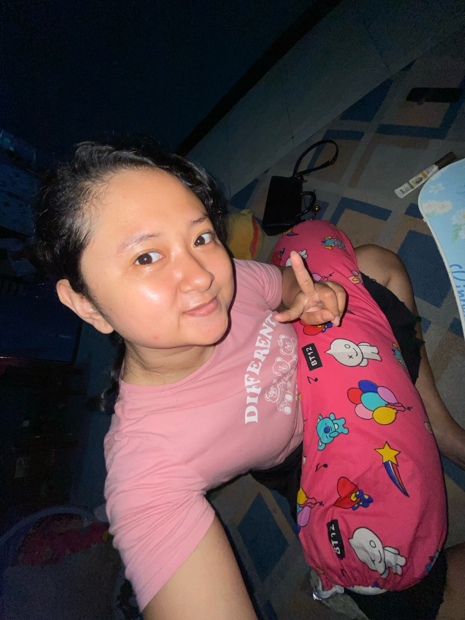 The image shows a young woman taking a selfie. She is wearing a pink t-shirt with the word "DIFFERENT" printed on it in white, and she is holding a pink pillow with cartoon characters on it. She is making a peace sign with her right hand. The background appears to be indoors, with a dark ceiling and a patterned floor. The scene is dimly lit. There are no specific details in the image that suggest a location in the city of Gelam, Indonesia.