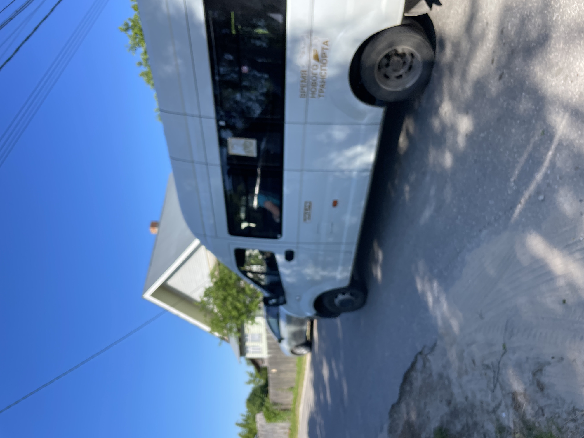 The image depicts a white minivan parked on a paved road in the city of Krasnye Baki, Russia, with a residential house and a wooden fence in the background. The minivan has dark tinted windows and features text on its side in Cyrillic script, which appears to read "ВРЕМЯ НОВОГО ТРАНСПОРТА" (Time of New Transport). A silver car is parked further down the road, to the left of the minivan. The sky is clear and blue, with power lines visible overhead. The ground beside the road is a mix of sand and vegetation. The scene appears to be a sunny day in a suburban or rural setting.