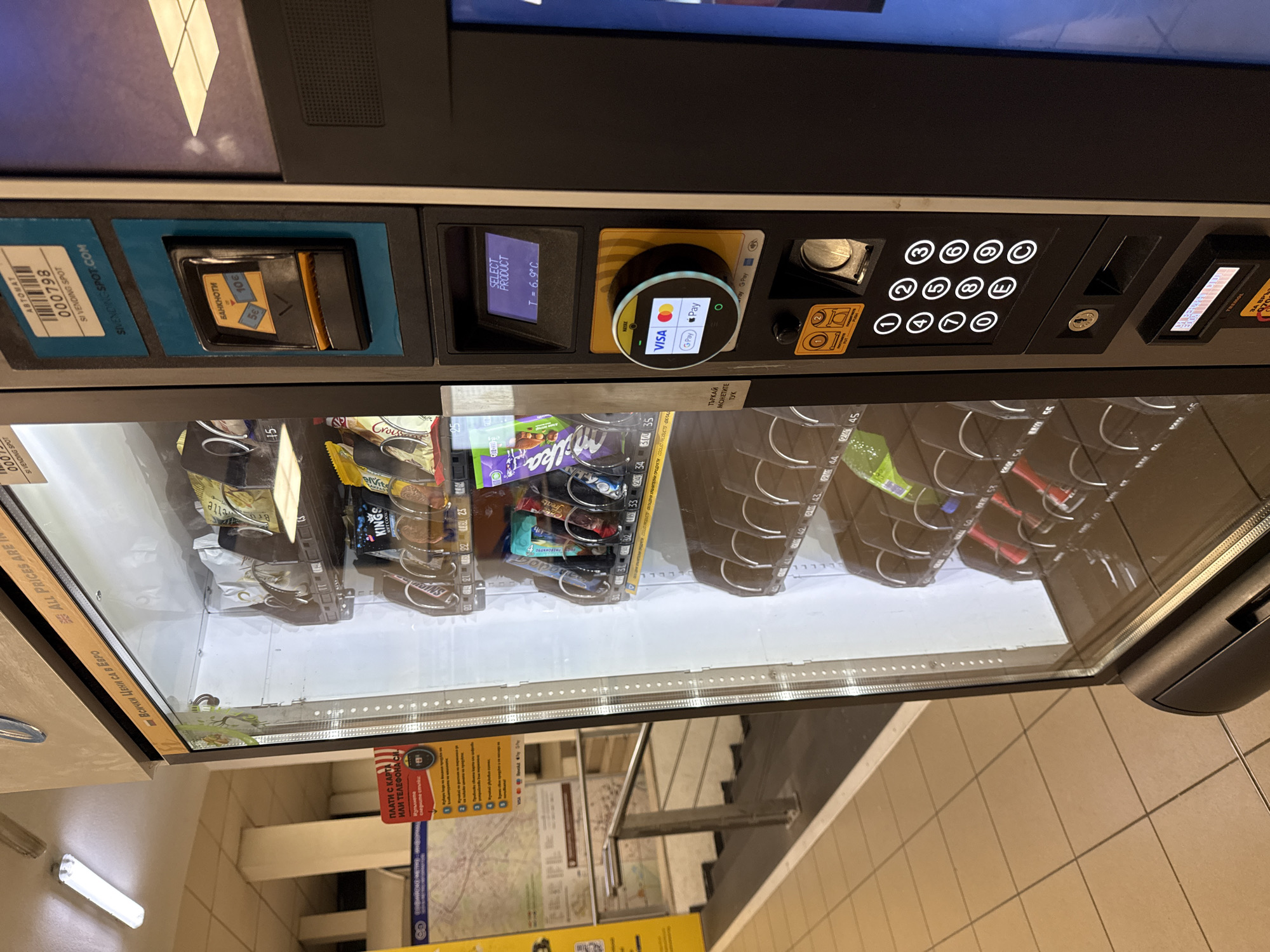 The image displays a modern vending machine, likely situated in an underground public space such as a metro station in Sofia, Bulgaria. The machine features a variety of payment and selection options on its control panel.
On the left side of the panel, a slot for inserting banknotes is visible, labeled "БАНКНОТИ" (BANKNOTES) with indications for 5€ and 10€ notes. A barcode and "SI VENDING SPOT" text are also present. Central to the panel is a small digital screen displaying "SELECT PRODUCT" and the internal temperature, "T = 6.9°C". Adjacent to this, a circular contactless payment terminal is prominent, showing logos for "VISA", "GPay", and "Pay", with a green light indicating it's ready for use. To the right, a numerical keypad (0-9, C, E), a coin slot, and a keyhole complete the interface.
Through the glass front of the machine, an assortment of snacks and beverages is visible, including Milka chocolate, Kings wafers, Croissant, Snickers, and various bottled drinks. Product codes are displayed on the shelves (e.g., 32, 33, 34, 35, 44, 45, 52, 53, 54, 55). Text on the machine states "ВСИЧКИ ЦЕНИ СА В ЕВРО" (ALL PRICES ARE IN EURO) and "COOLED PRODUCTS".
The surroundings include a light-colored tiled floor. Reflections in the machine's glass reveal stairs and a handrail, reinforcing the likely setting of a public transport hub. A hanging sign with Cyrillic text, "ПЛАТИ С КАРТА ИЛИ ТЕЛЕФОНА СИ" (PAY WITH CARD OR PHONE), further details the available payment methods. No individuals are present in the image.