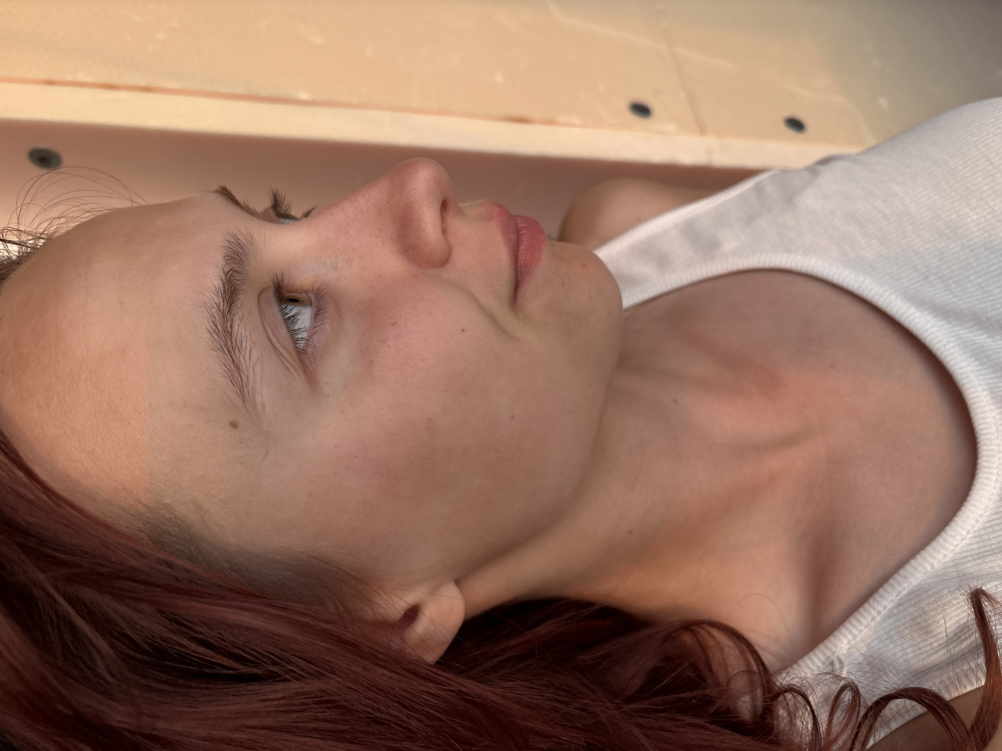 A close-up, profile shot shows a young Caucasian woman with reddish-brown hair and fair skin, facing right. She is wearing a white tank top. Her left ear is visible, and her neck and shoulder are also in view. The background appears to be a light-colored wall with visible hardware. The lighting is warm and suggests an indoor setting, possibly with natural light. The image does not provide any contextual information about a specific location in Прикамский, Russia.