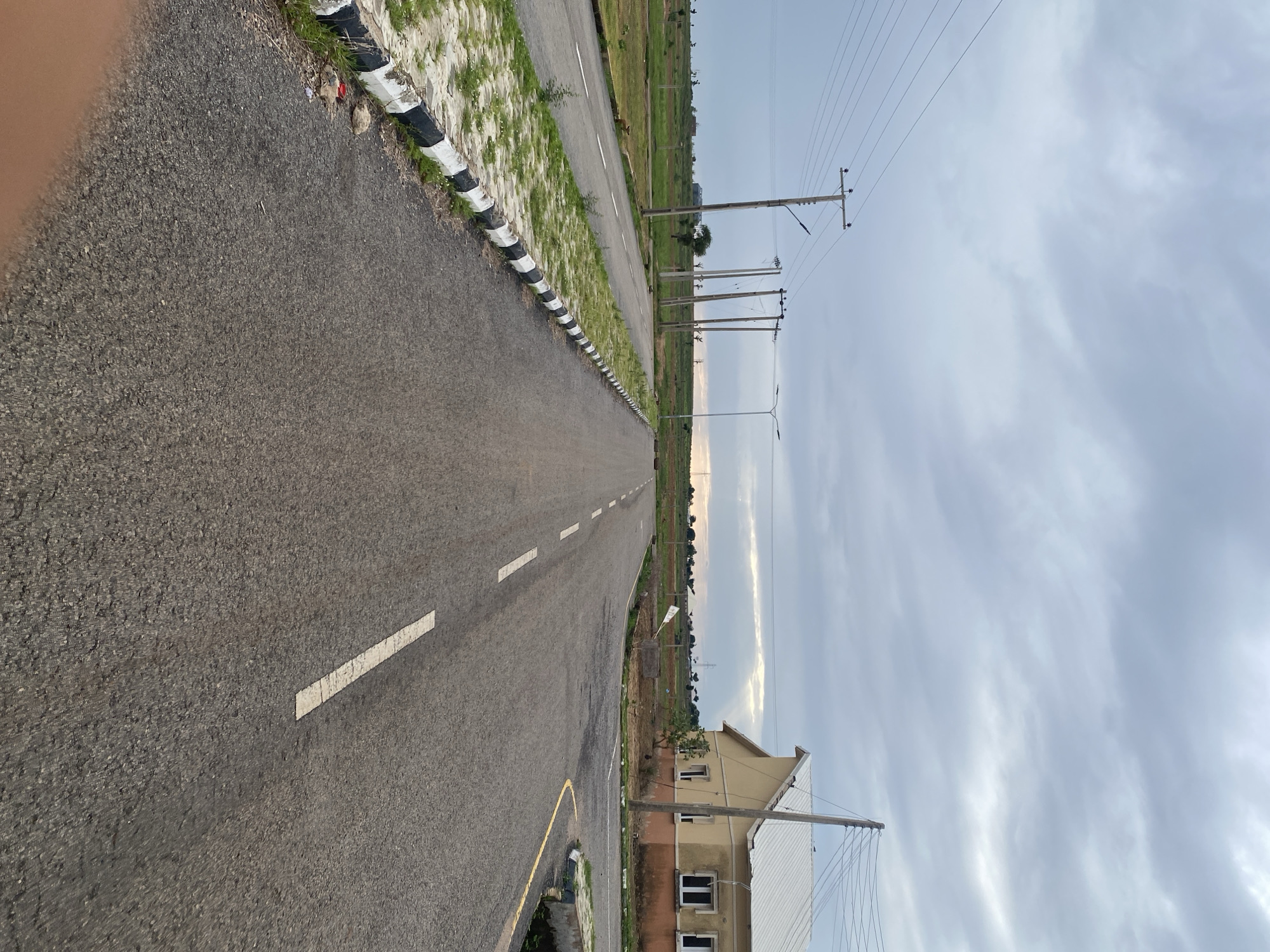 The image depicts an empty asphalt road with white dashed markings, curving towards a distant horizon. The road is bordered on the left by a strip of grass and a black and white striped curb. In the distance, on the right side of the road, a two-story building with a cream-colored facade and a white roof is visible, alongside a utility pole with electrical wires. The scene is set under an overcast sky, suggesting a cloudy or possibly a pre-rainy day. The presence of utility poles and the architecture of the building hint at a developed or developing residential area in Nigeria, likely in Bulamare given the location context. There are no people or specific activities visible in the image, giving it a tranquil, perhaps slightly isolated feel. The lighting suggests it is either early morning or late afternoon.