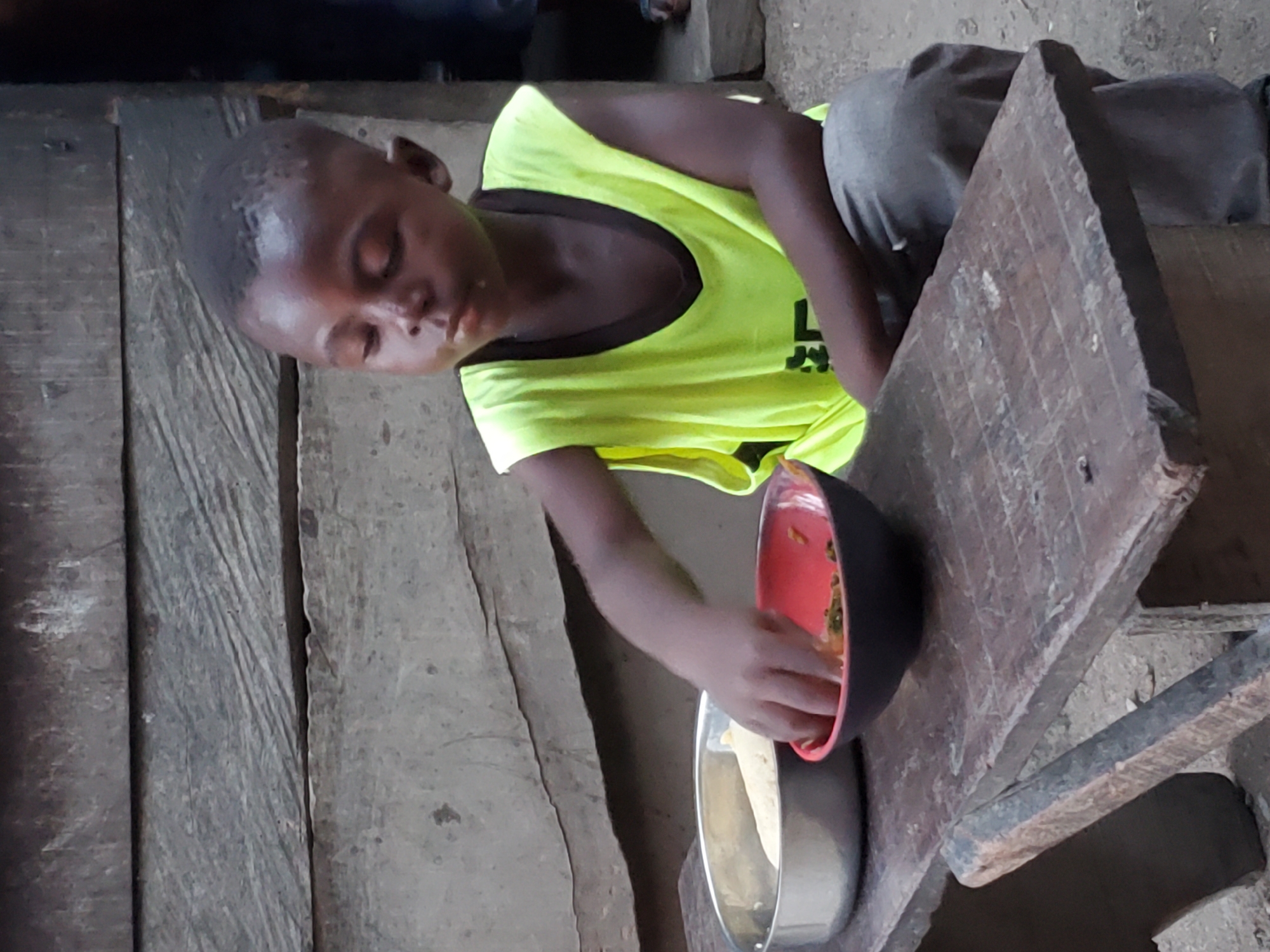 A young Black child is depicted eating a meal in a simple, rustic setting. The child, appearing to be in early childhood, is wearing a bright neon yellow t-shirt with a dark trim around the neck and grey trousers. Their hair is very short, and their eyes are closed or intently focused downwards on the food.
The child's right hand is immersed in a red and black bowl, which contains a greenish, stew-like mixture, possibly vegetables or a local delicacy. Beside it, on a rough wooden surface, is another silver-colored bowl, though its contents are less distinct. This wooden surface appears to be a makeshift table or bench.
The background consists of dark, weathered wooden planks, suggesting an interior space or a sheltered area. The wood shows signs of age and wear. The overall scene conveys a moment of quiet focus as the child consumes their meal. There is faint, illegible text visible on the front of the child's yellow t-shirt. The context of Mararaba, Nigeria, aligns with the visual cues of a daily life scene in a modest environment.
